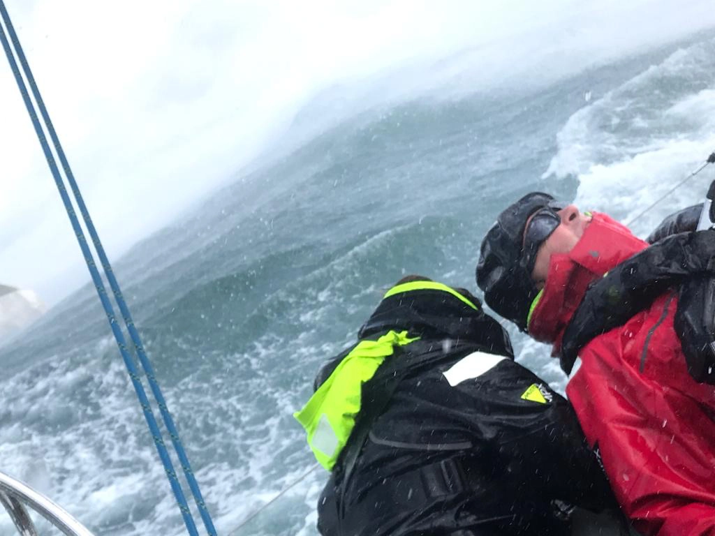 Two people in waterproof jackets and caps on a sailboat in rough, choppy sea waters with overcast skies.