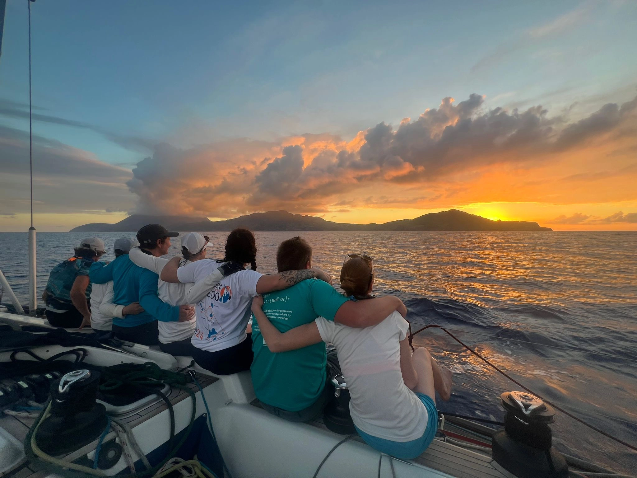 Group of seven people sitting on a boat, arm in arm, watching a vibrant sunset over the ocean and distant mountainous islands.
