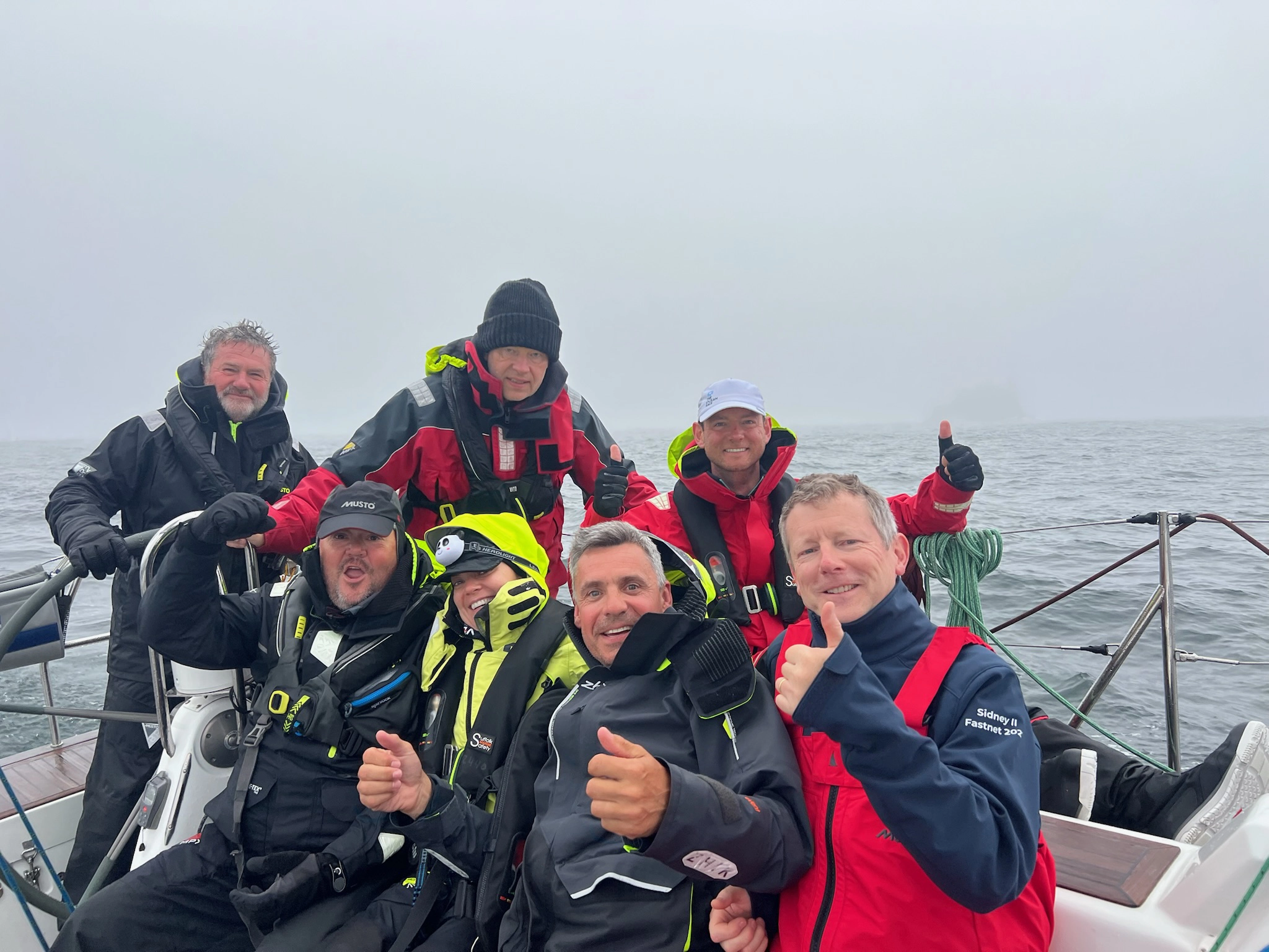 Six sailors on a boat in protective gear, smiling and giving thumbs up on a foggy sea.
