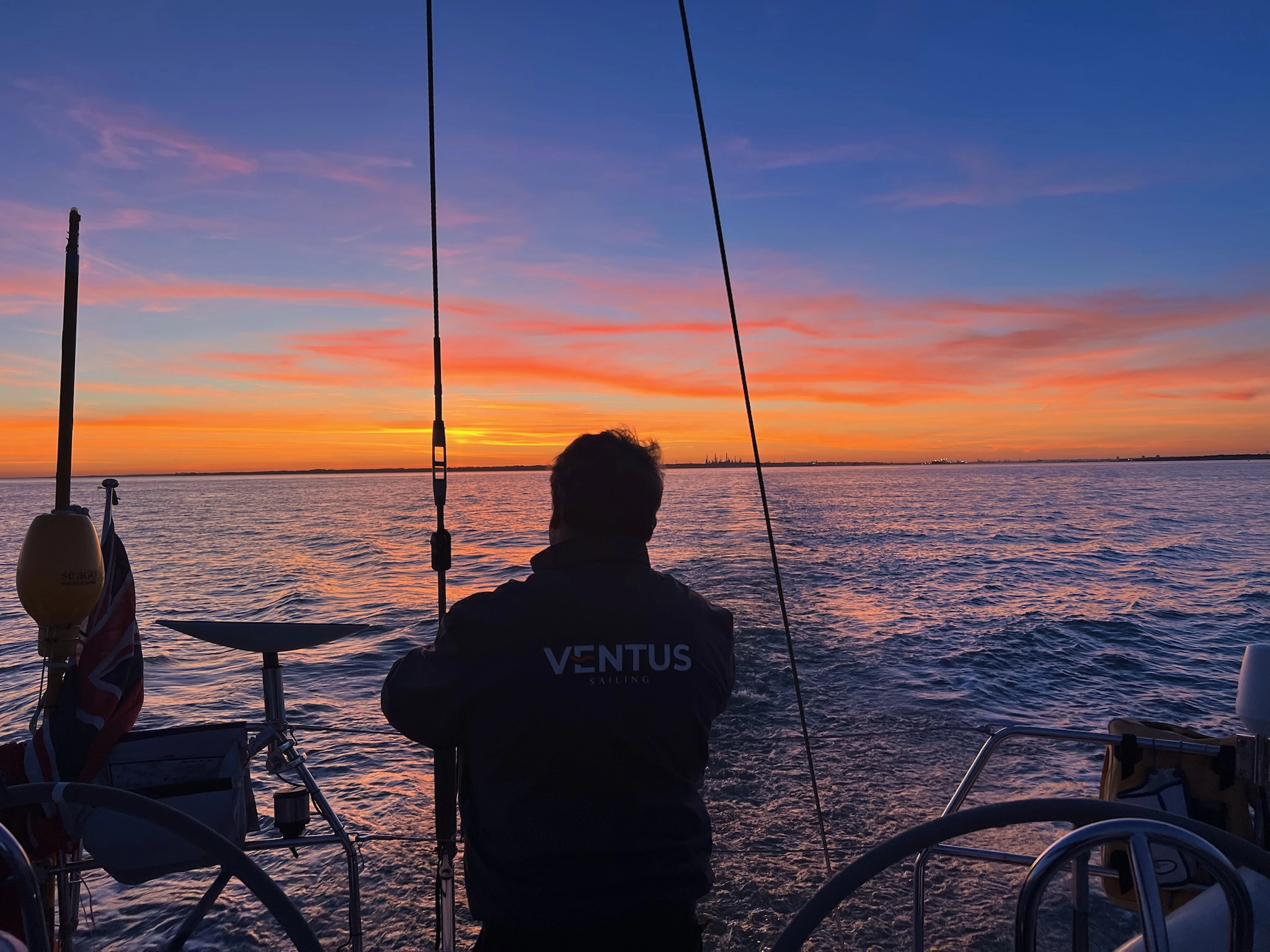 Person on a sailboat at sunset with an orange and purple sky over calm water.