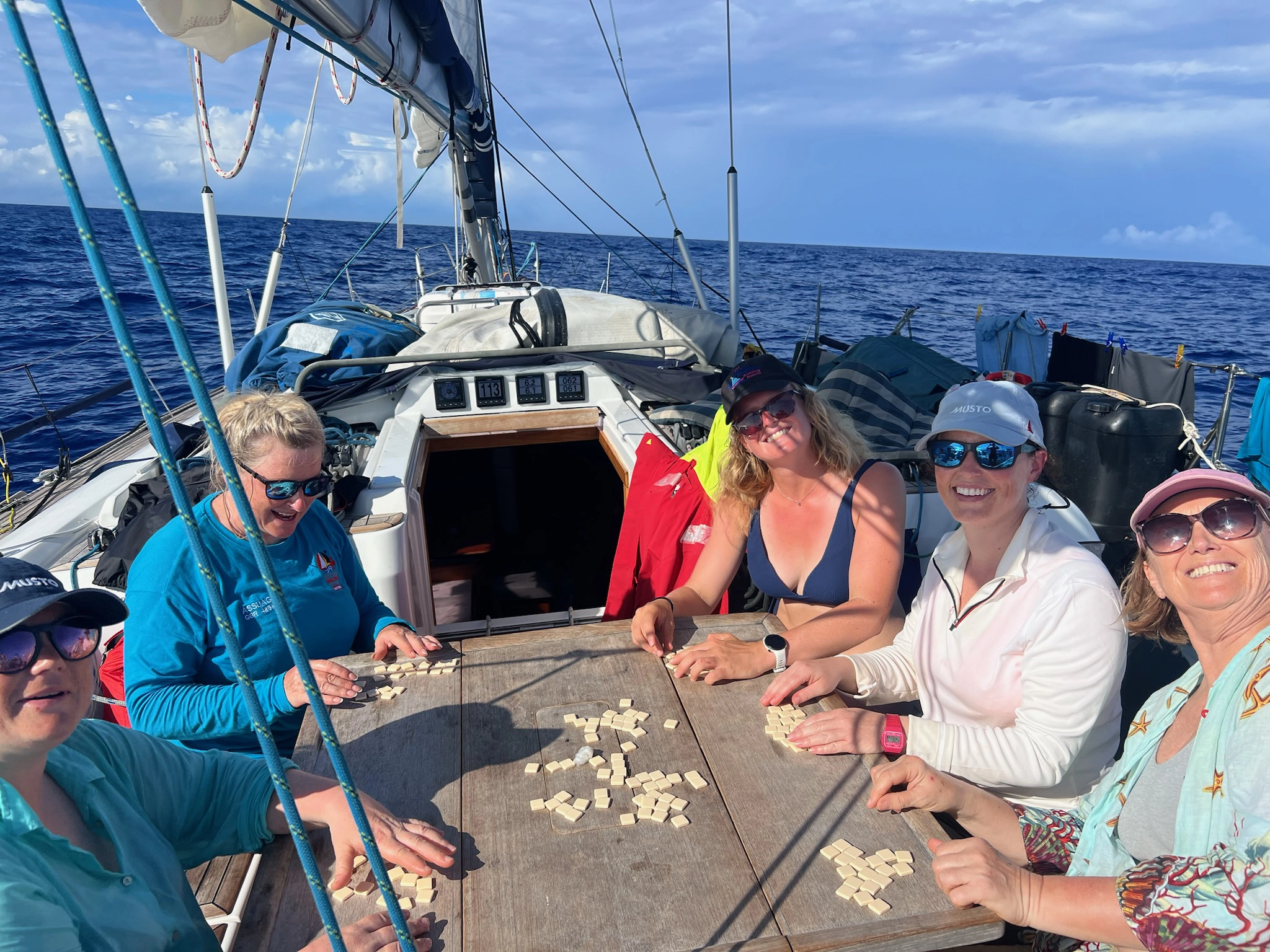 Five women smiling and playing a tile game around a wooden table on a sailboat in the open ocean.
