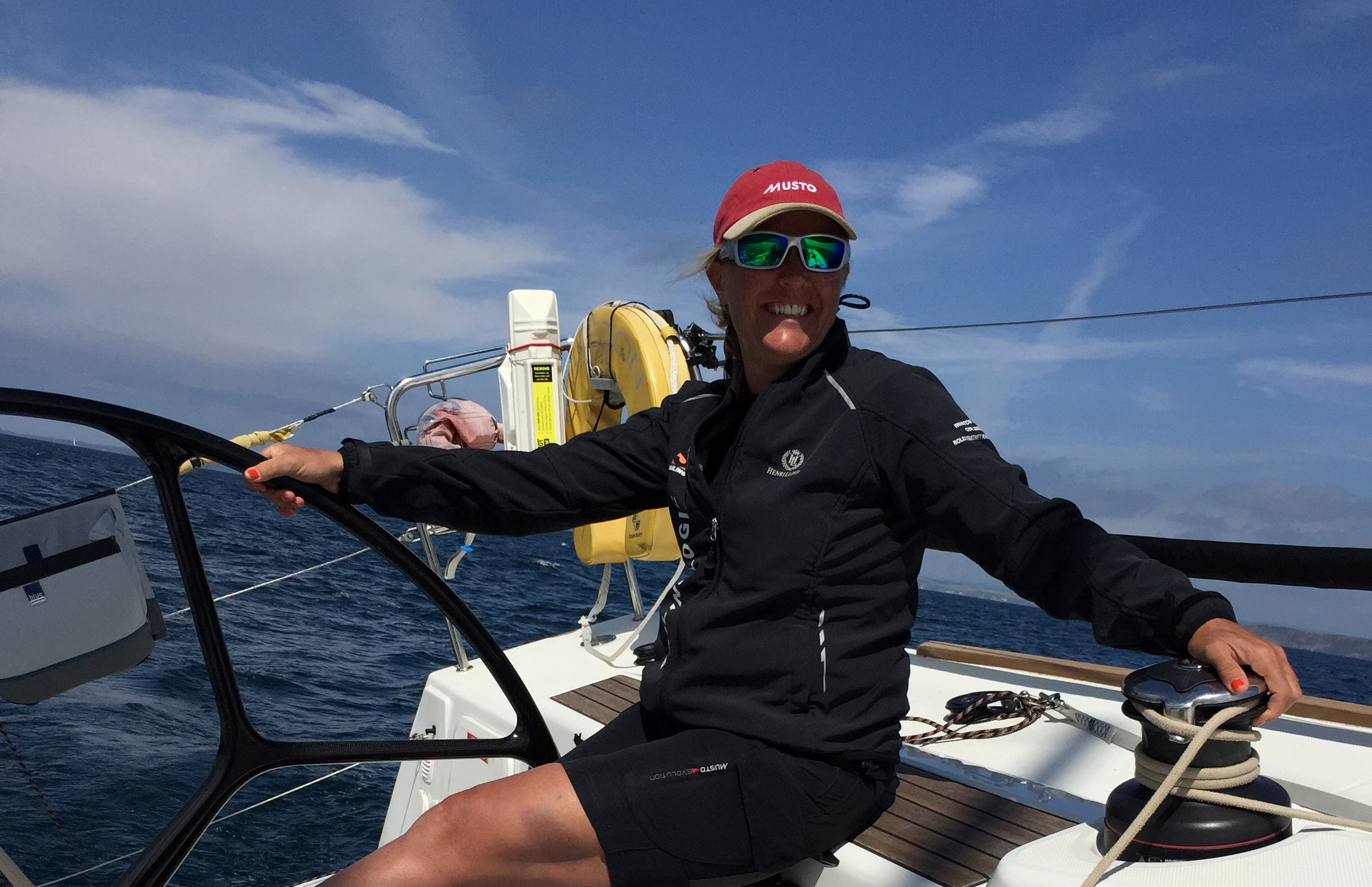 Smiling woman wearing sunglasses and a red Musto cap steering a sailboat on a sunny day with a blue sky and ocean background.