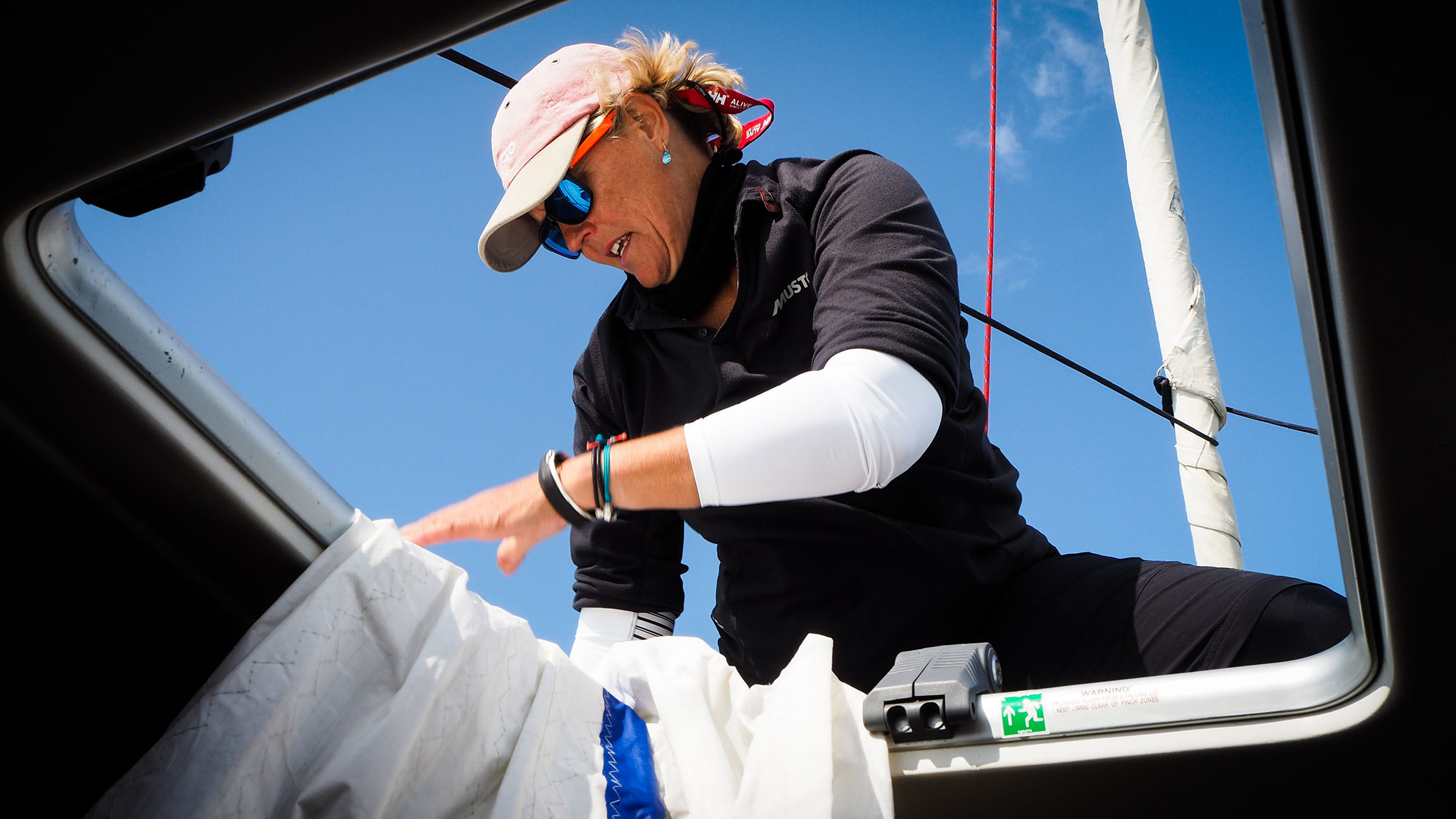 Person wearing a cap, sunglasses, and black clothing adjusting a white sail on a boat under a clear blue sky.