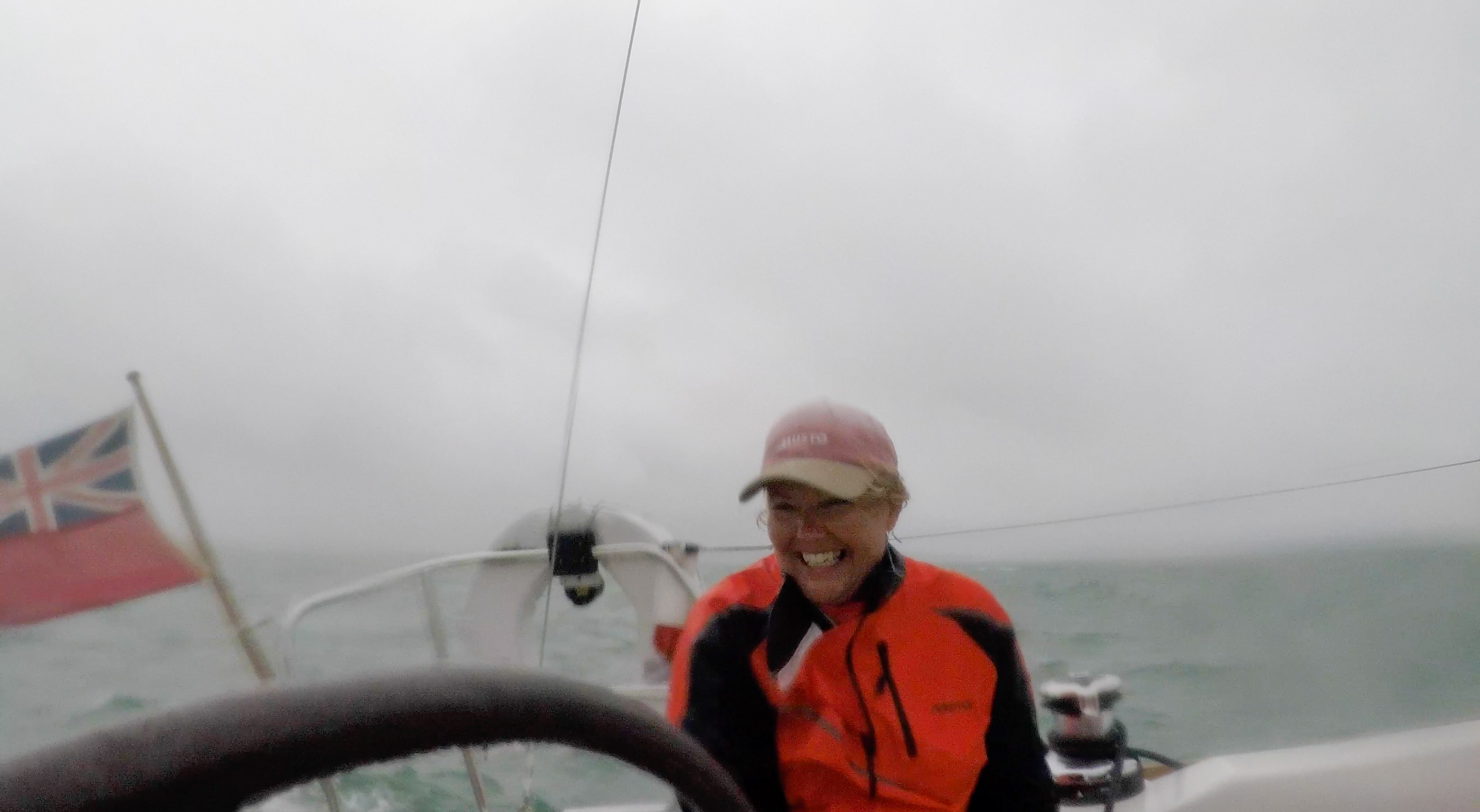 Person in an orange jacket and pink cap smiling on a sailboat with a red ensign flag in the background on a gray, cloudy day.
