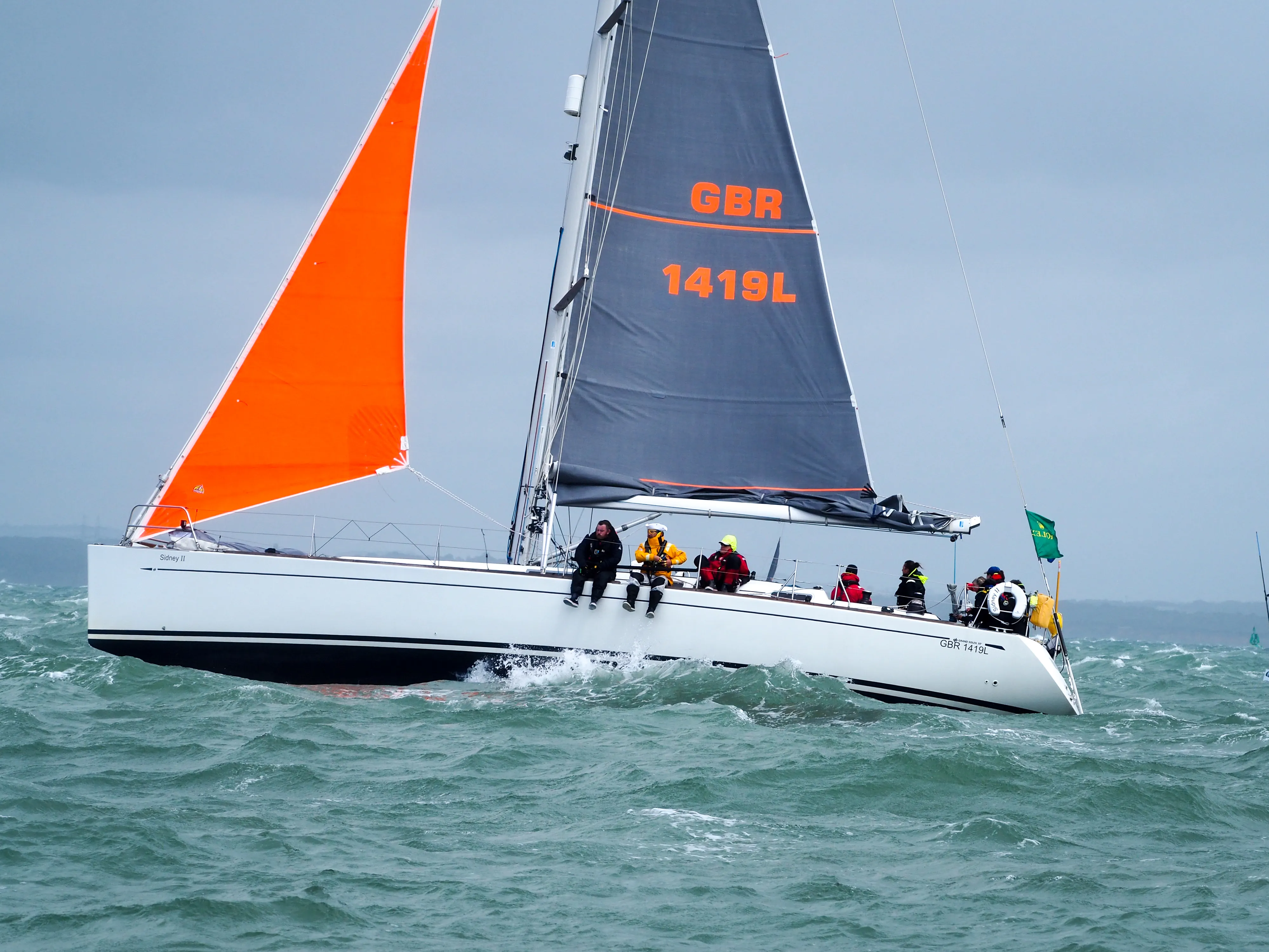White sailboat with bright orange and black sails sailing on choppy green waters under a cloudy sky, with crew members on board.