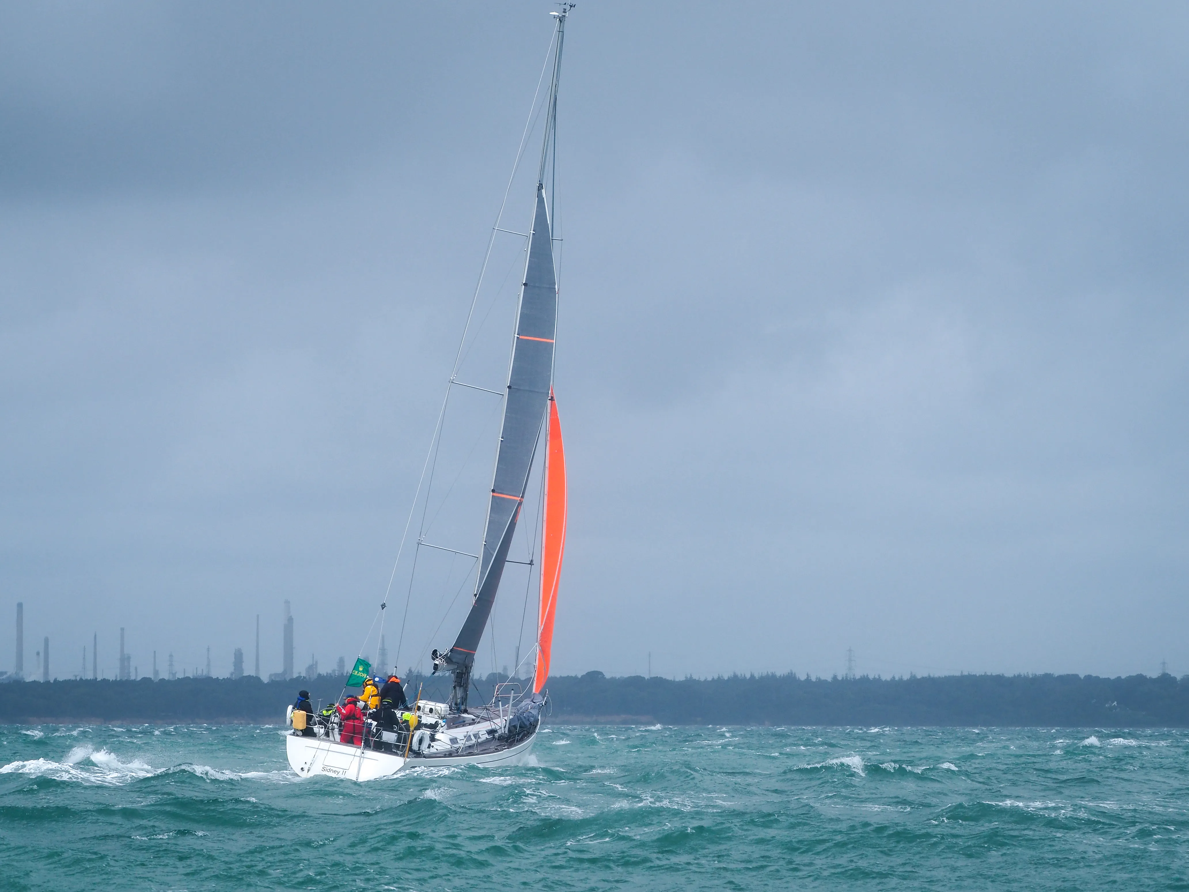 Sailboat with a red and gray sail in choppy waters under cloudy sky with industrial buildings on distant shore.
