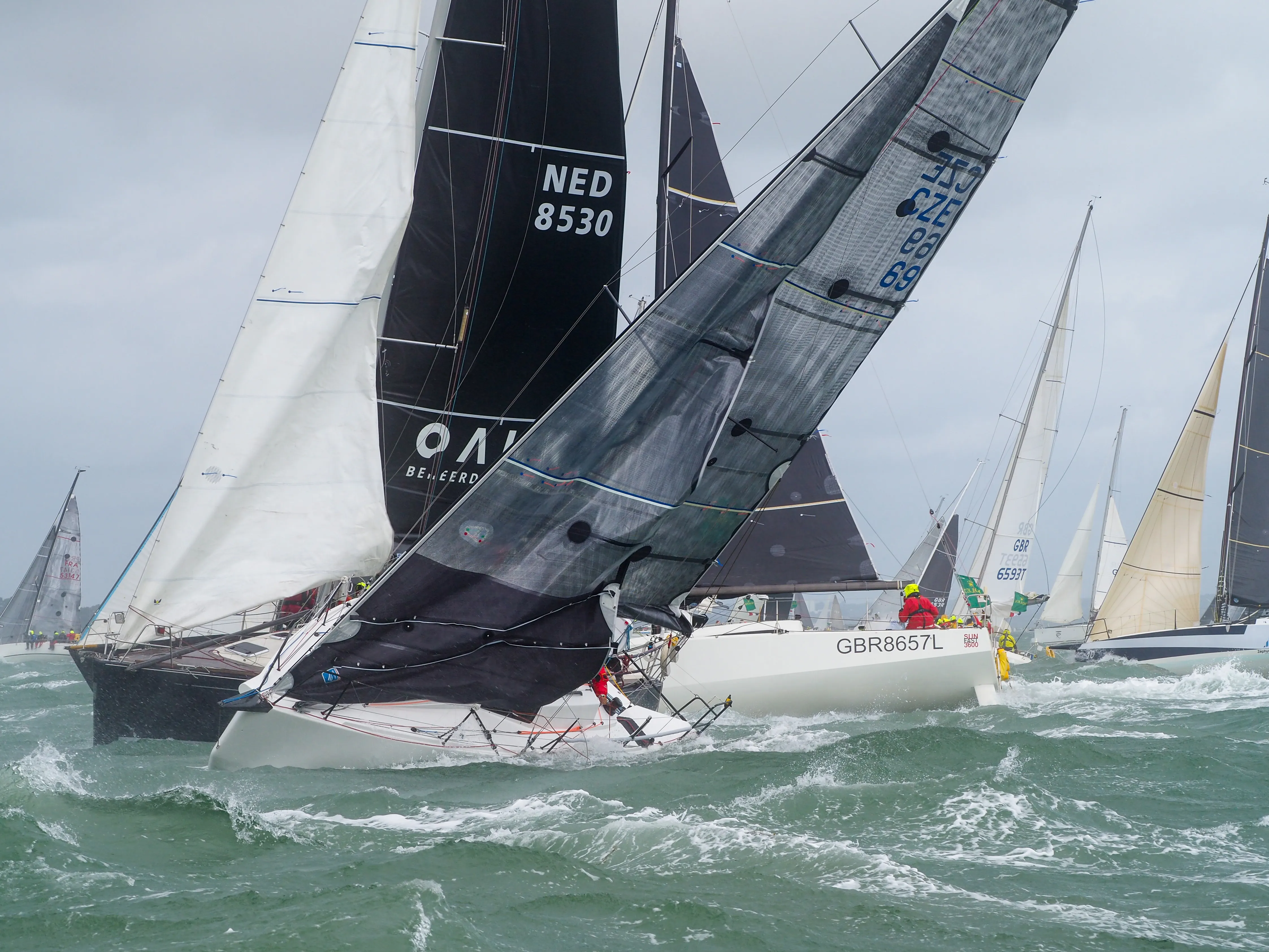 Several sailboats with colorful sails racing on choppy green water under a cloudy sky.