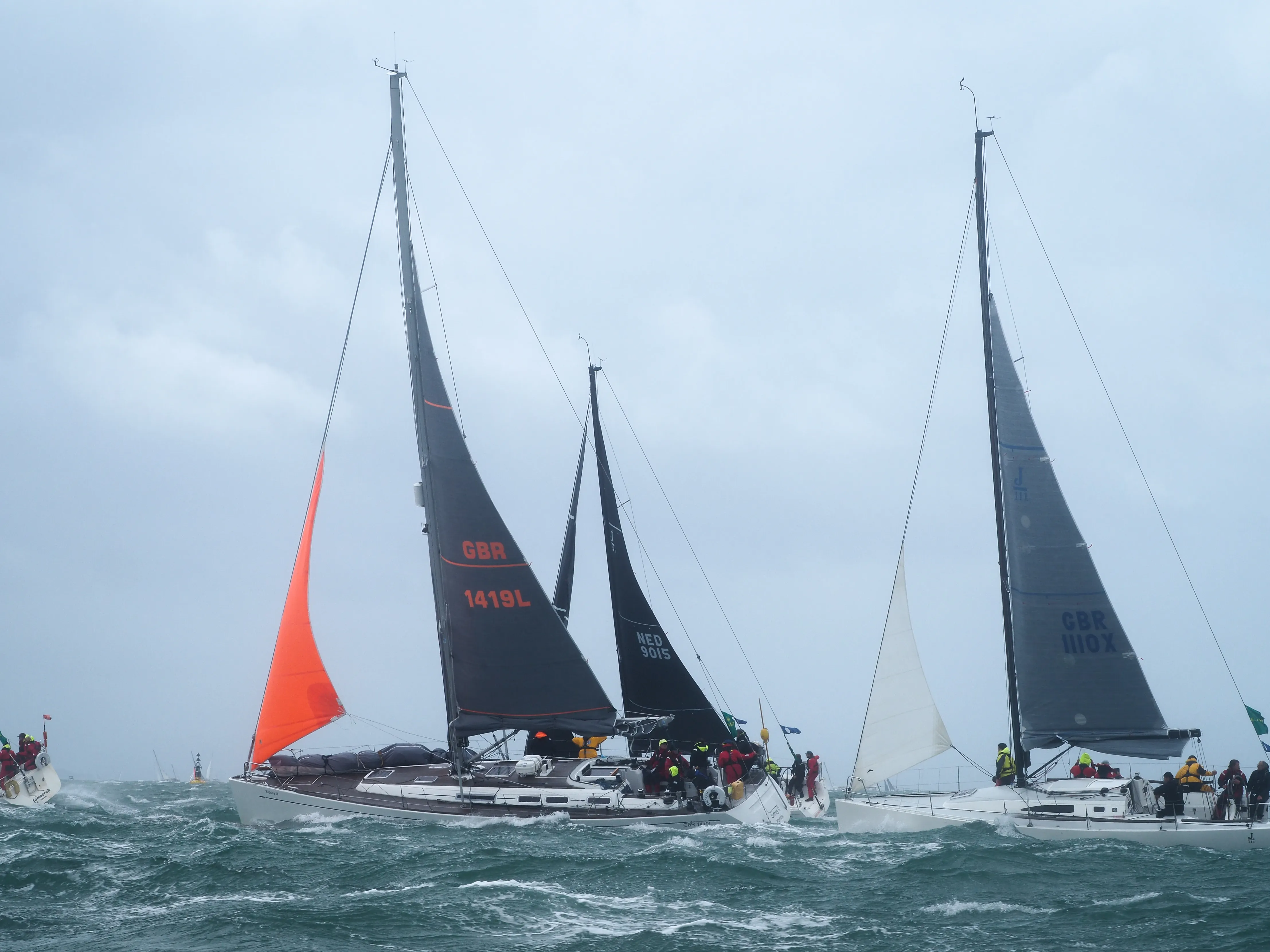 Group of sailboats with crews navigating rough waters under a cloudy sky during a sailing race.
