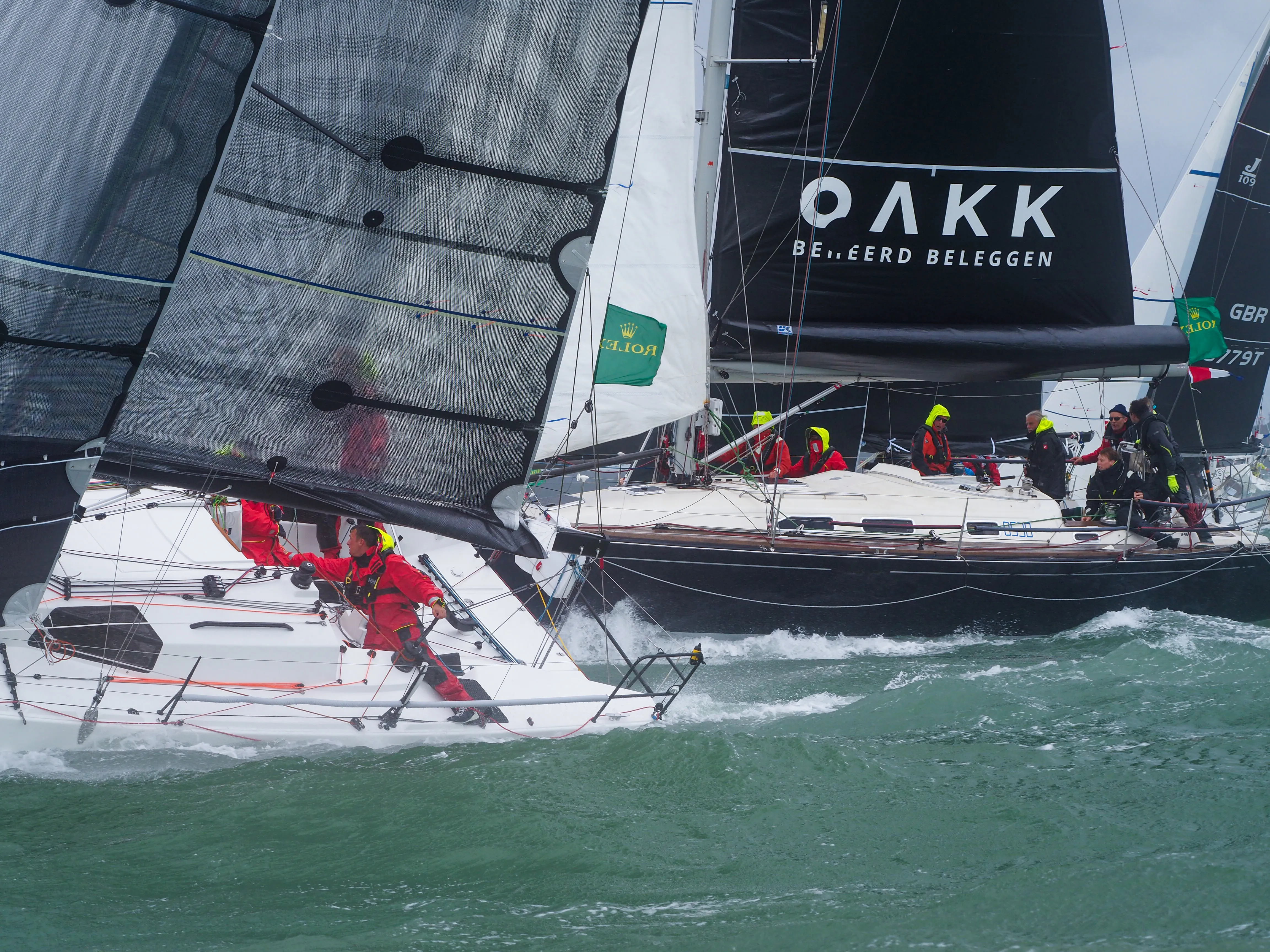 Several sailors in bright gear active on two competing sailboats with black and white sails in choppy waters.