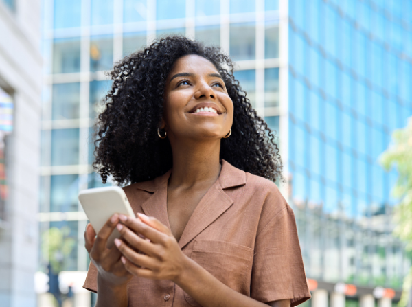 Happy professional African American business woman holding smartphone looking away with cellphone in hands outdoors, young lady customer using mobile technology apps on cell phone standing in city