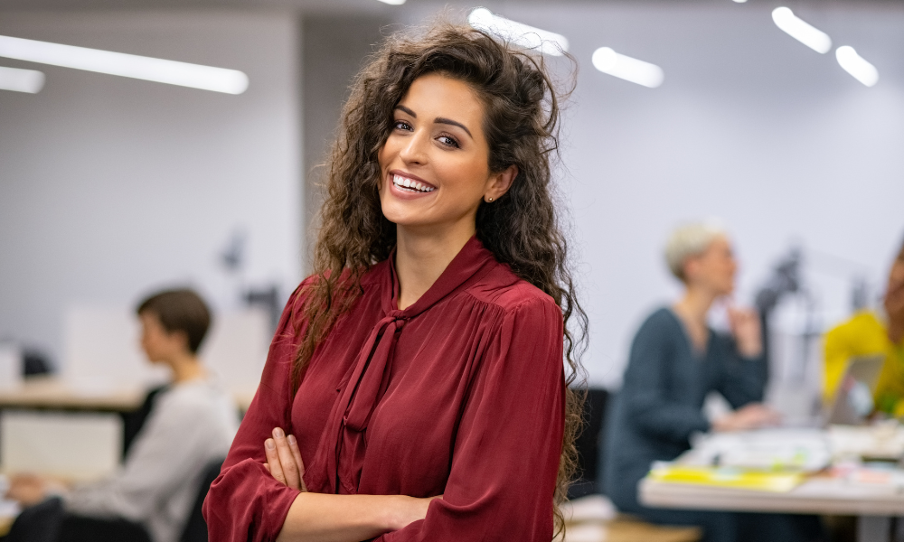 Professional woman with curly dark hair smiling confidently in a modern office environment. She is wearing a burgundy blouse and standing with her arms crossed while coworkers can be seen working in the background.