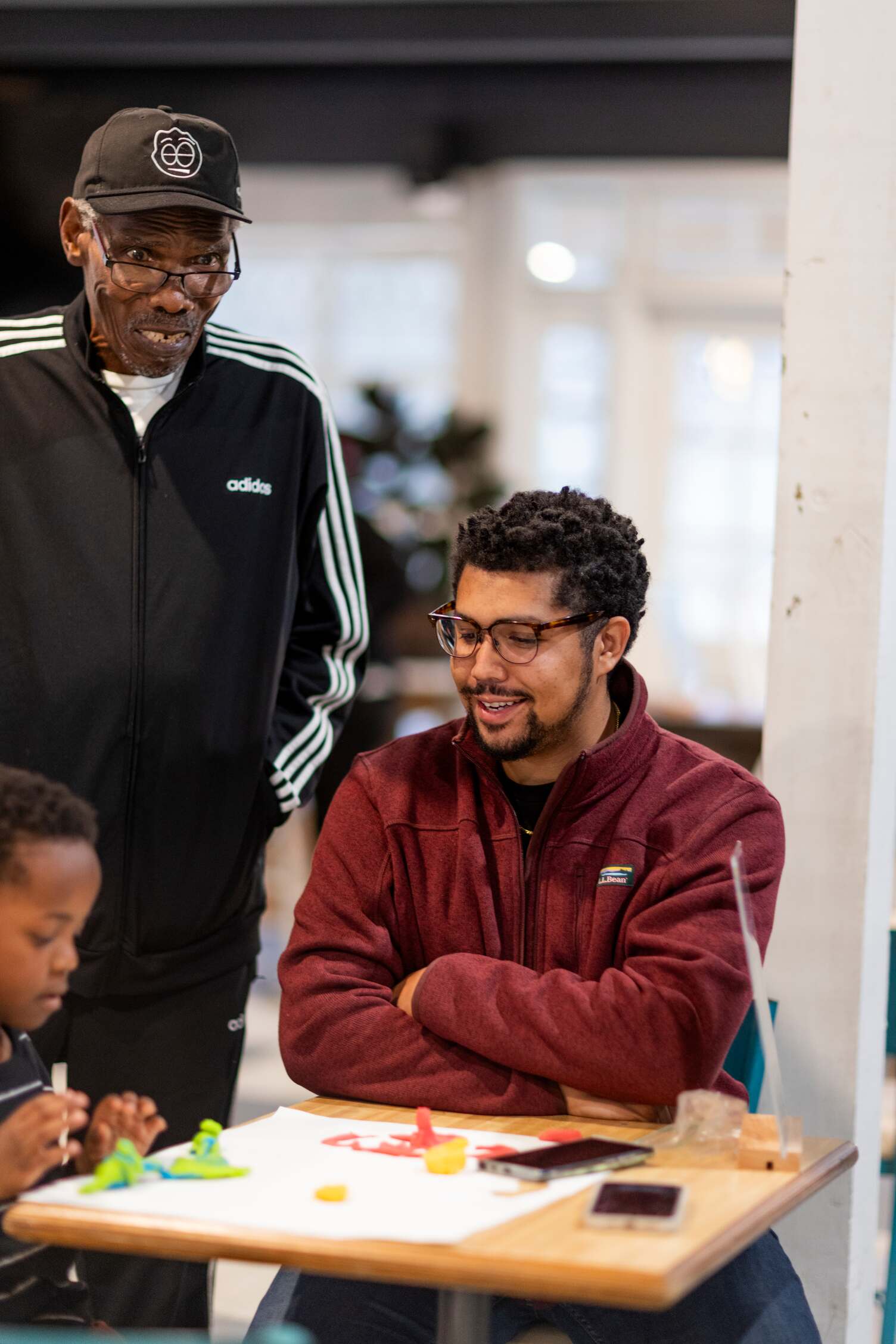 A man in a track suit standing next to a man sitting at table playing a game with a child