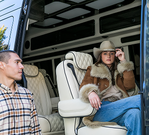 A woman in a fur-lined jacket and hat sits inside a vehicle while a man in a plaid shirt stands near the open door.