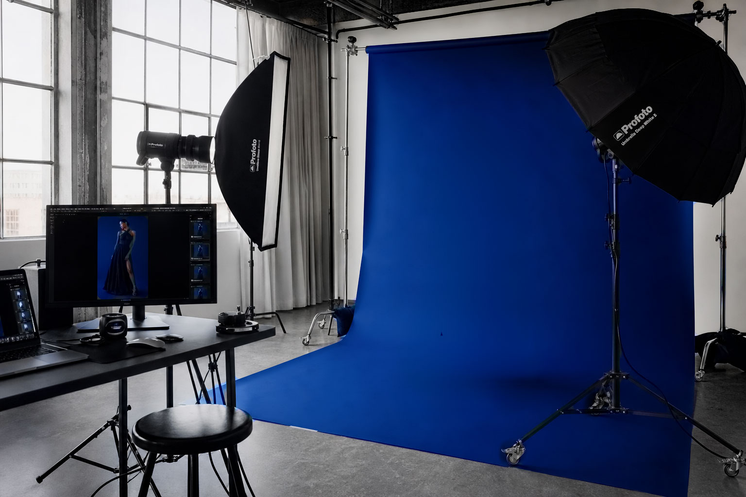 Photography studio with blue backdrop, lighting equipment, computer monitor showing model in a dark dress.