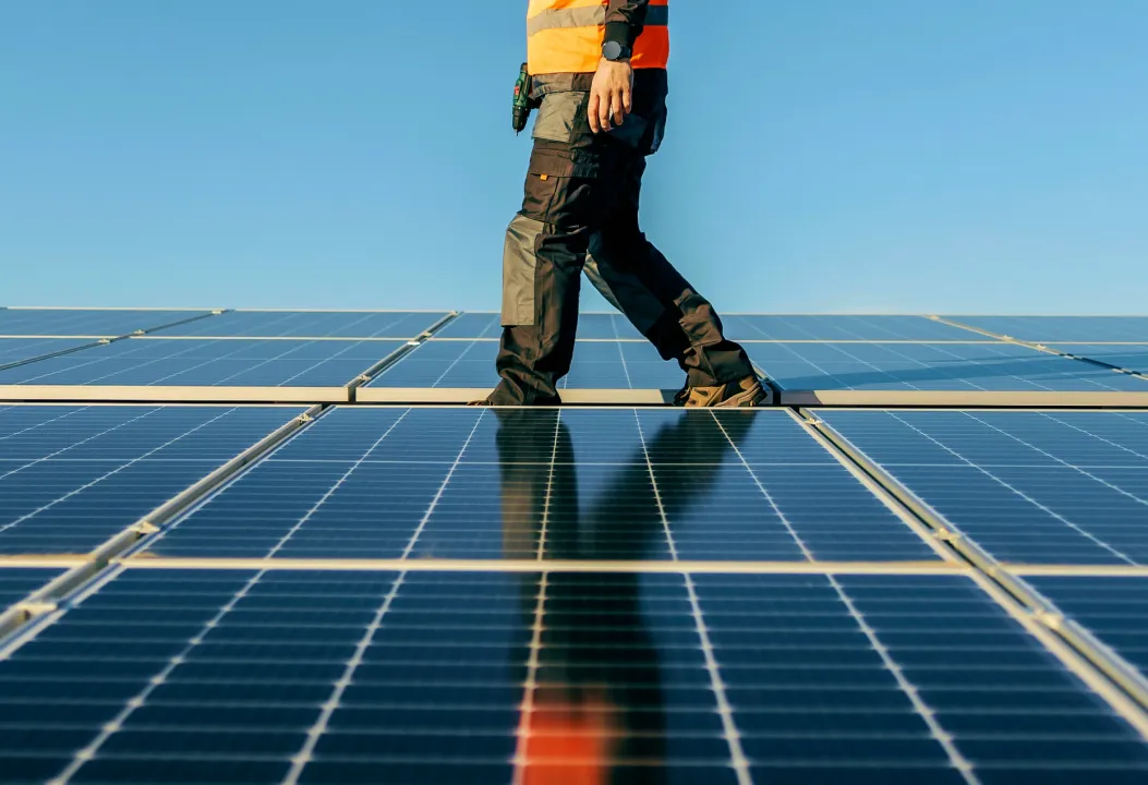 A technician wearing a high-visibility vest walks across a large installation of solar panels under a clear blue sky.