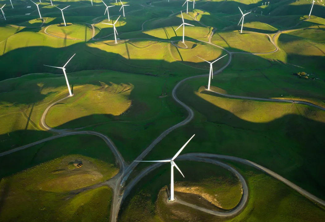 An aerial view of a large wind farm with numerous wind turbines spread across rolling green hills.
