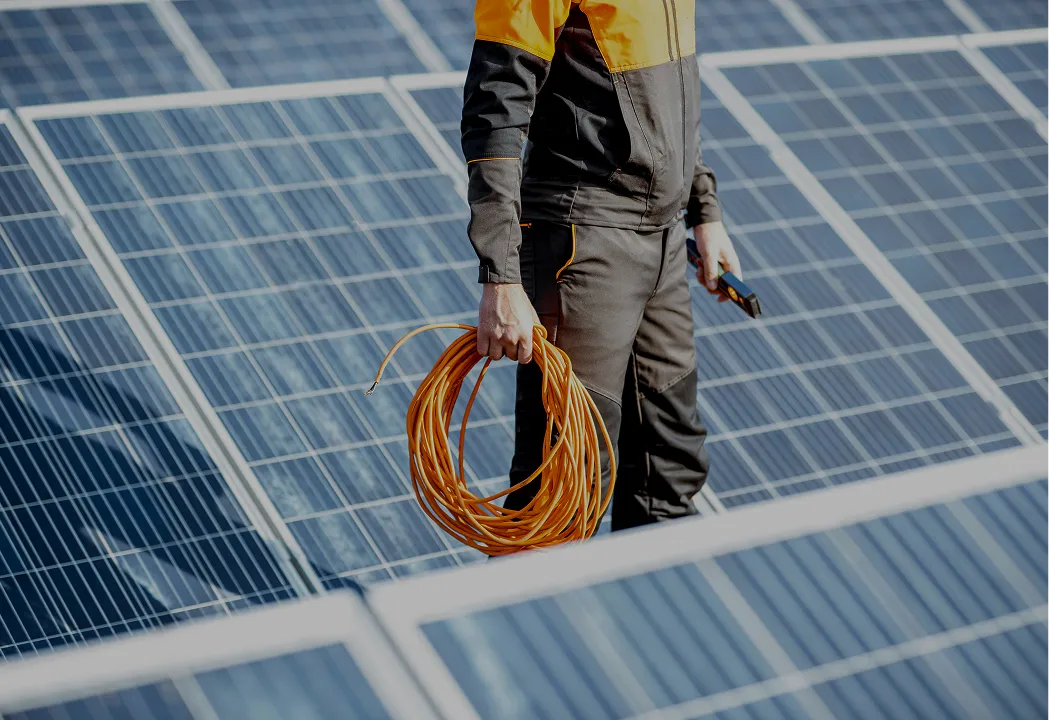 A technician stands on a large array of solar panels, holding a coil of orange electrical cable and tools.