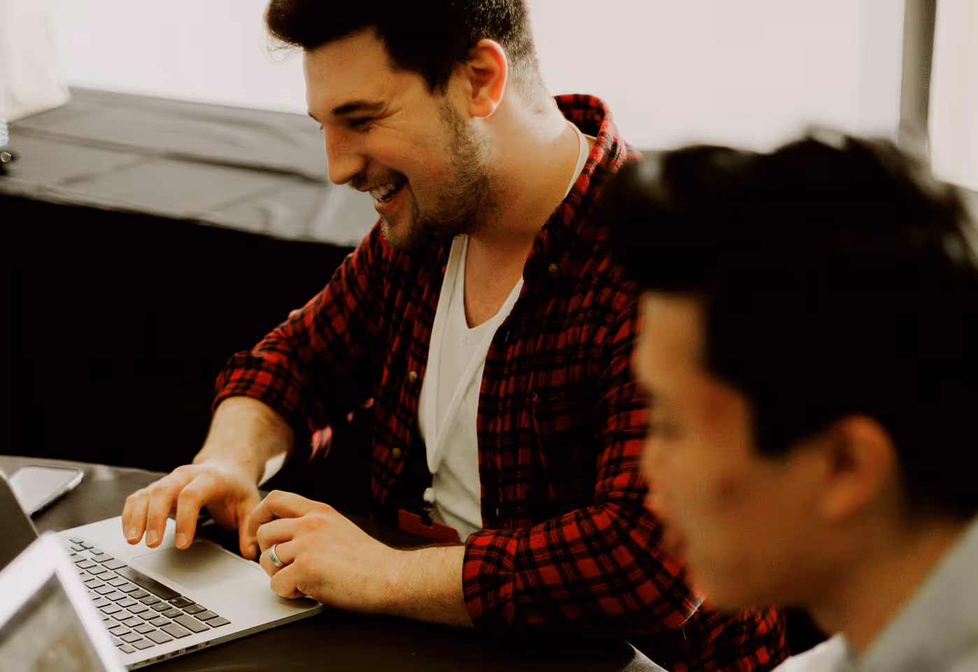 Two men working on laptops, one smiling and wearing a red plaid shirt.