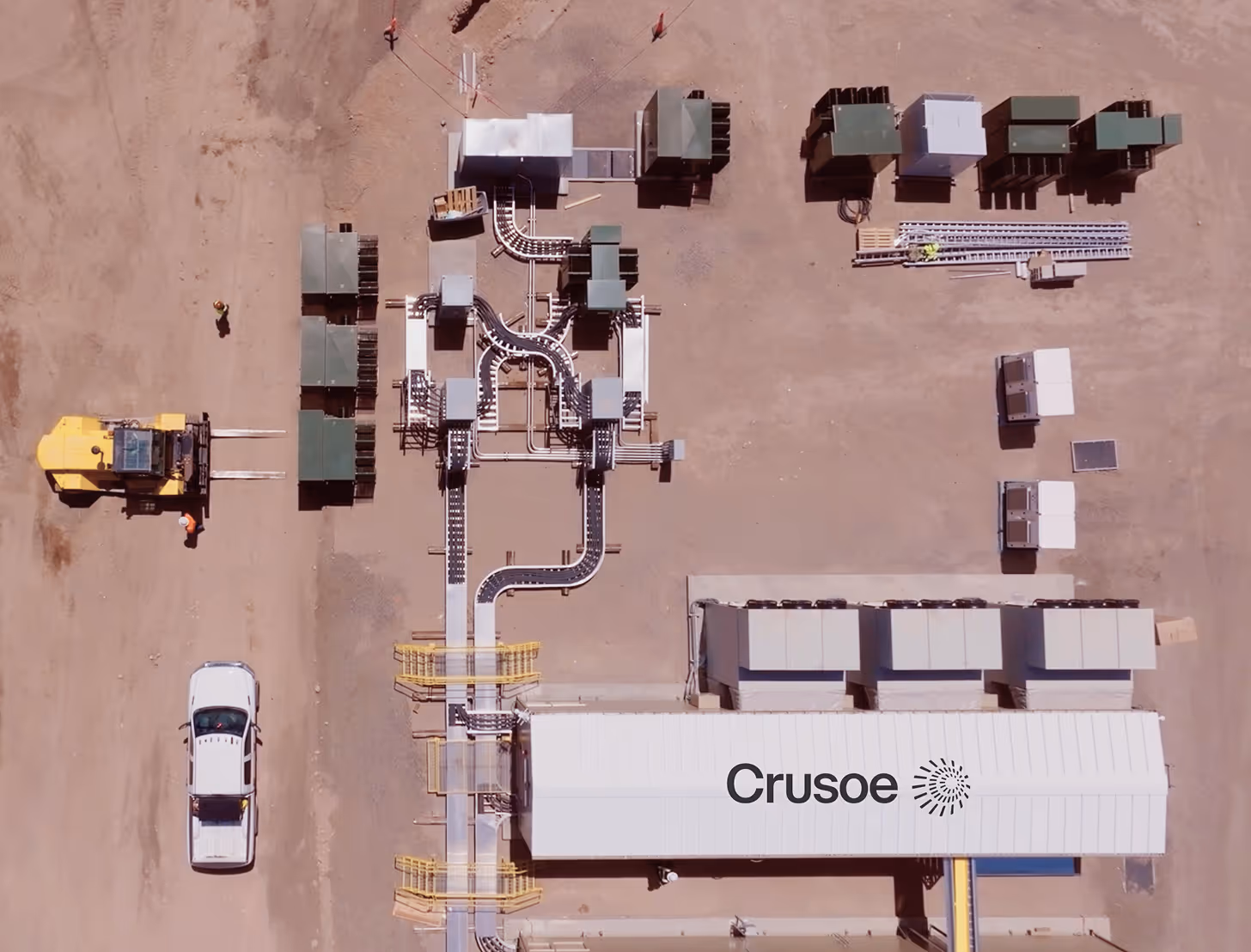Aerial view of an industrial site with white and yellow buildings, conveyor belts, a white pickup truck, and a yellow forklift on a dirt ground.
