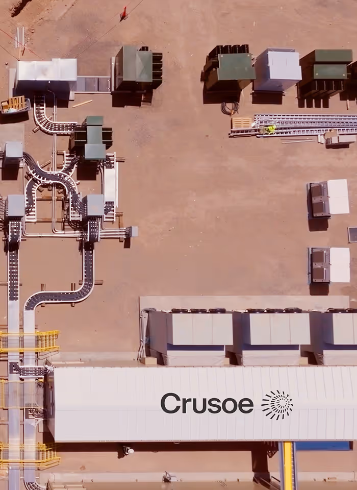 Aerial view of an industrial site with buildings, conveyor belts, and equipment on a sandy ground, featuring the Crusoe company logo.