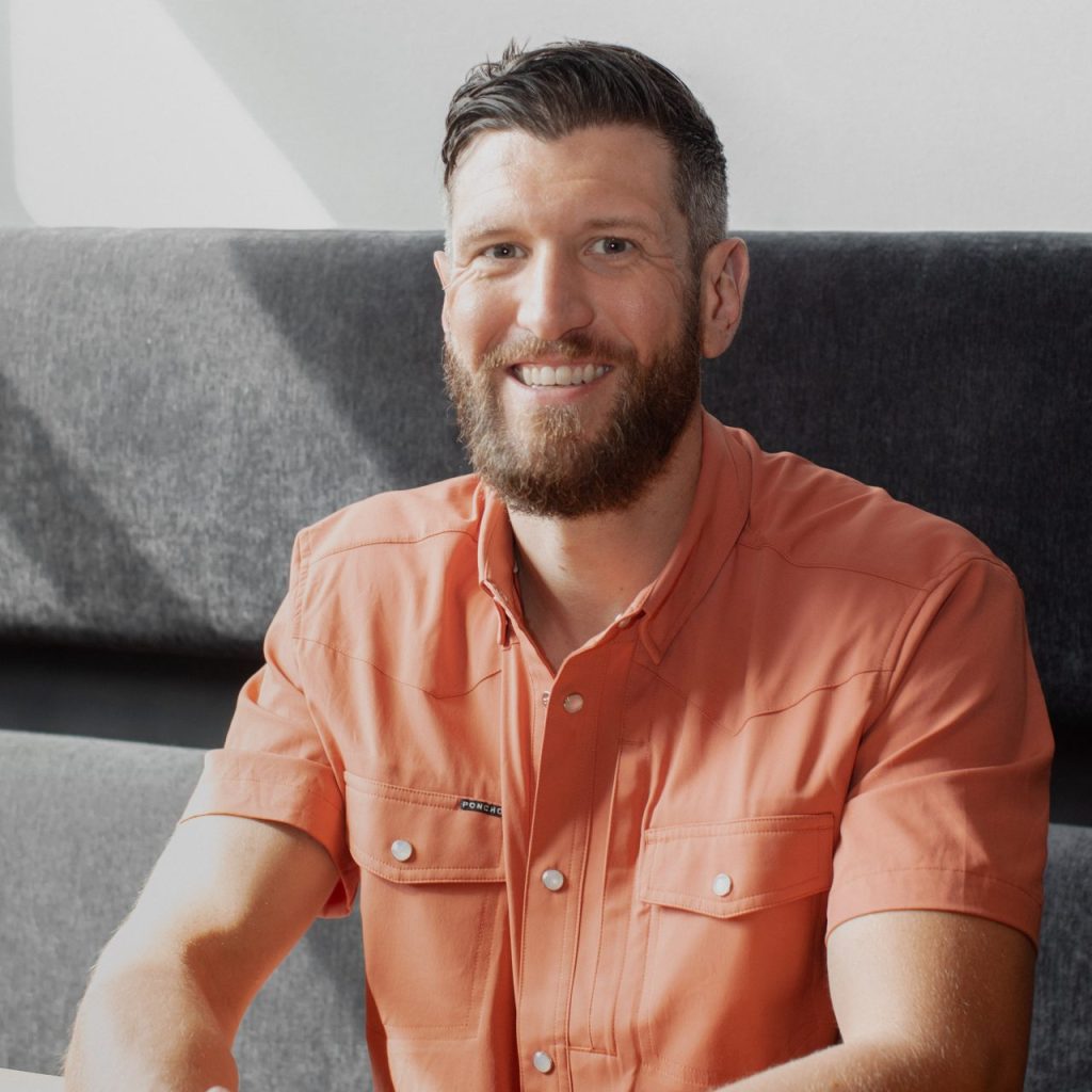 Smiling bearded man in an orange short-sleeve button-up shirt sitting indoors on a gray couch.