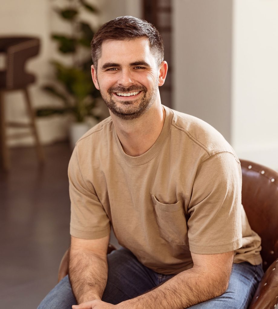 Smiling man with dark hair and beard wearing tan shirt and jeans sitting on brown chair indoors.