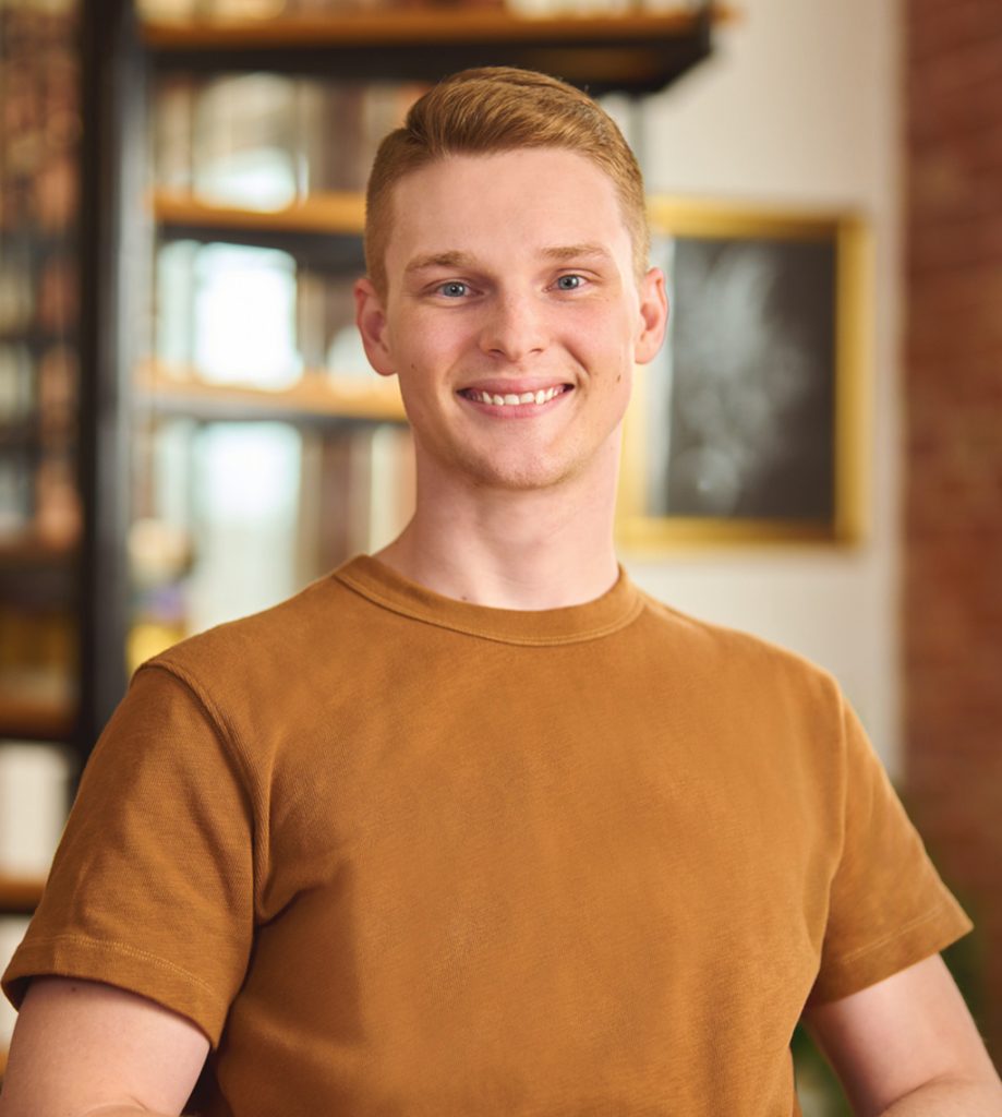 Smiling young man with short blond hair wearing a brown t-shirt in a softly lit indoor space.