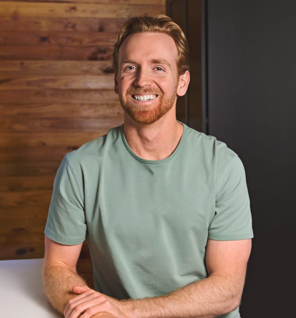 Smiling red-haired man in a light green t-shirt sitting with hands clasped indoors.