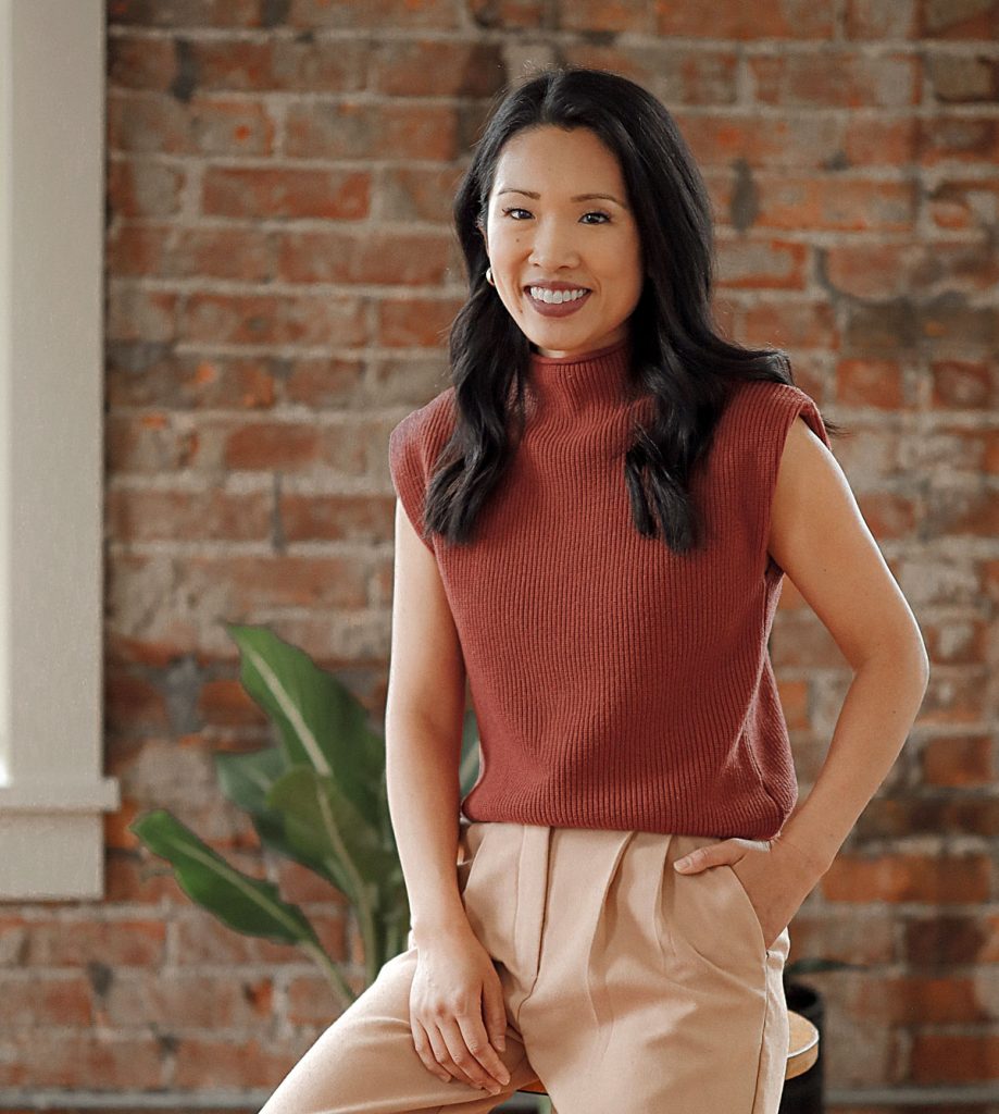 Smiling woman with dark hair wearing a sleeveless rust sweater and beige pants seated by a brick wall.