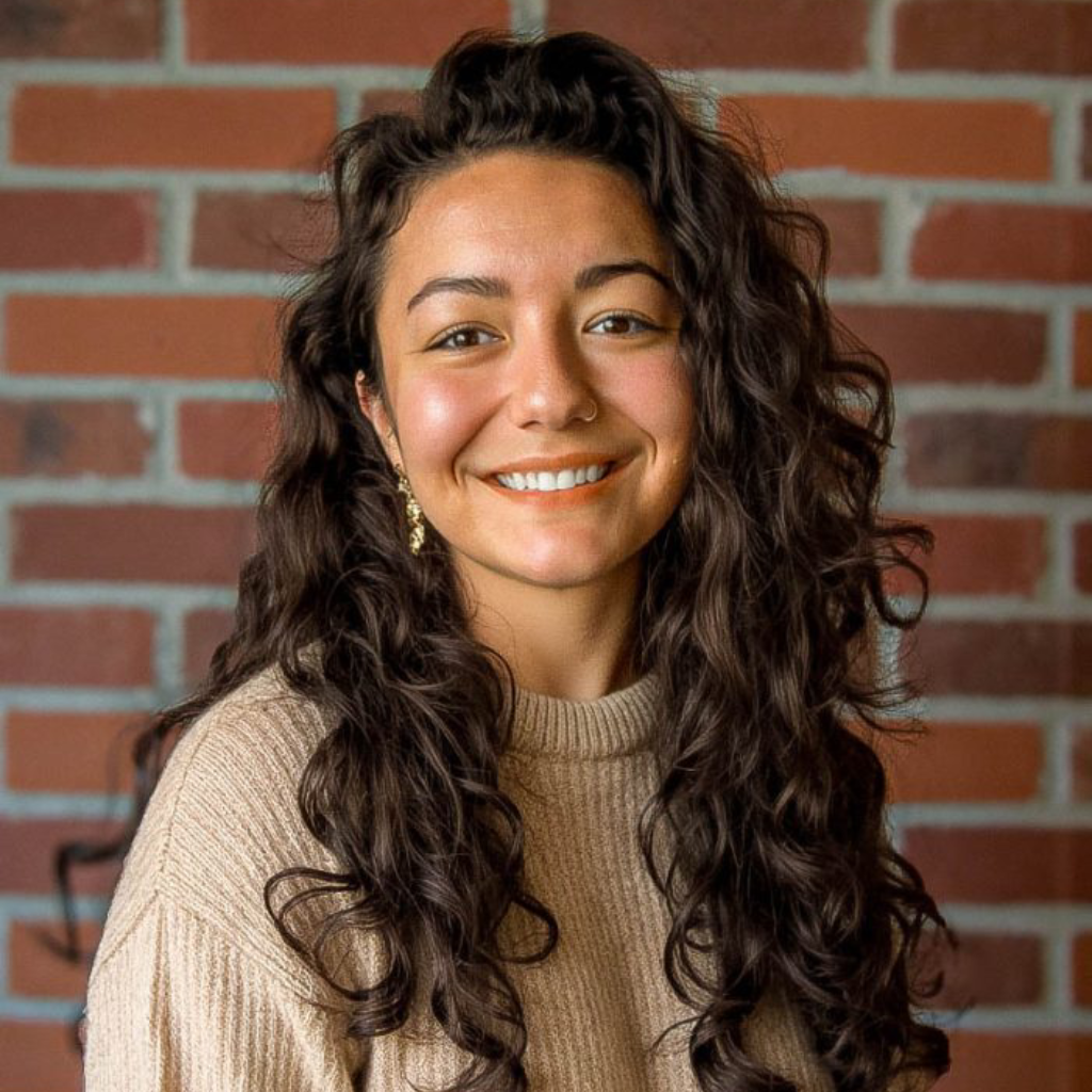 Smiling woman with long curly dark hair wearing a beige sweater in front of a brick wall.