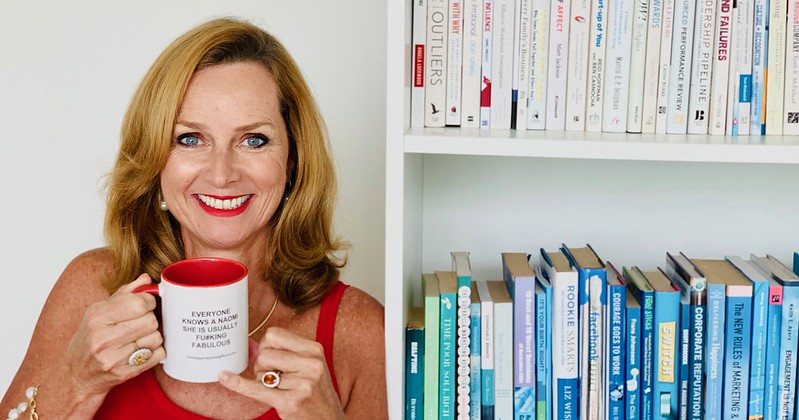 Naomi Simson smiling with a red mug in front of a bookshelf of business books