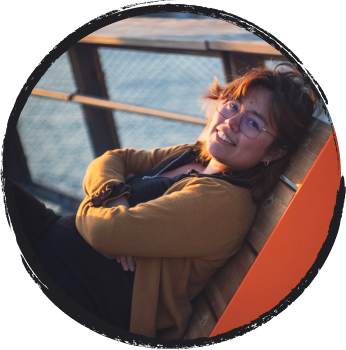 An about me button with a picture of Lidia: She smiles, wearing a mustard yellow cardigan and reclining on a bright orange park bench.