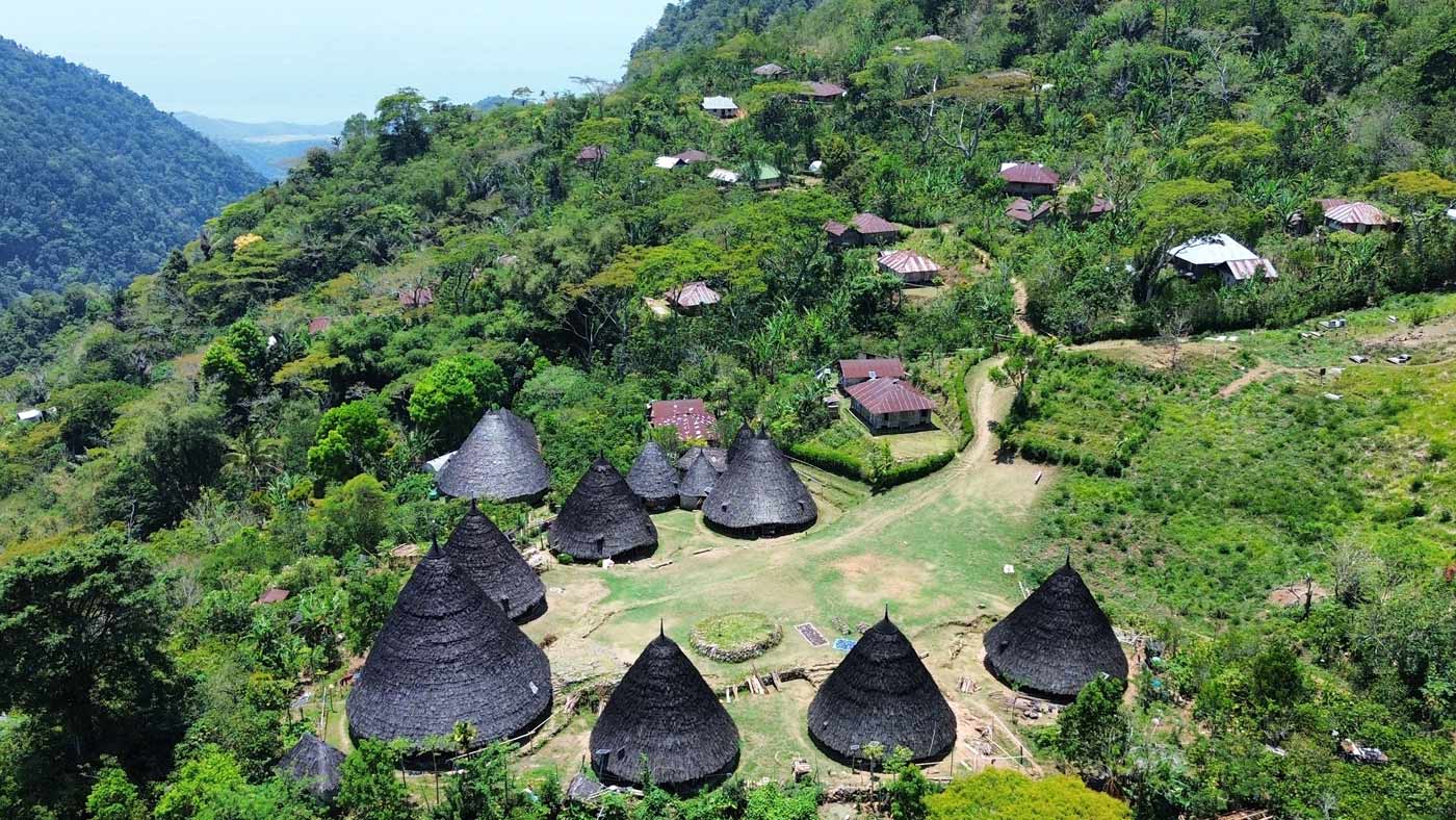 An aerial view of the Wae Rebo village, showing the group of unique, conical Mbaru Niang houses arranged in a circle in the foreground. In the background, other village houses and the surrounding lush green mountains are visible.