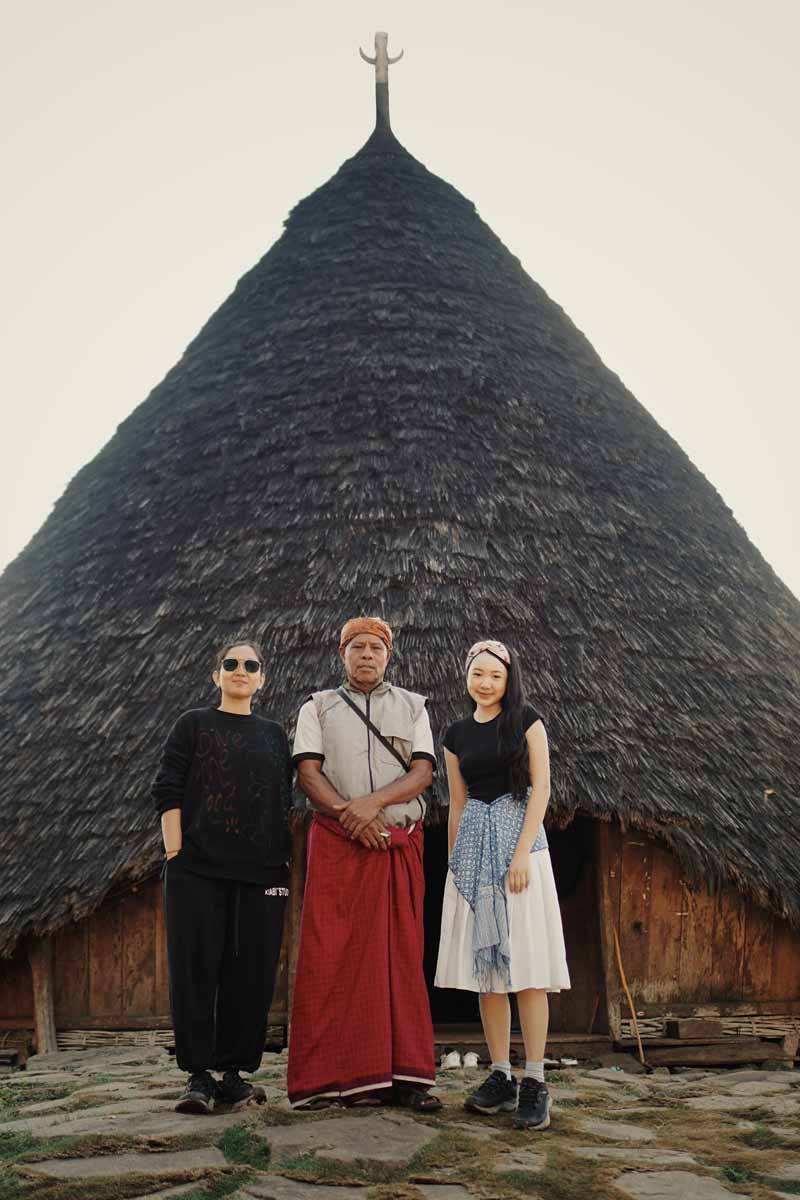 A group of three people standing in front of a traditional Wae Rebo house. The house has a tall, conical, thatched roof. The person in the center wears a traditional red wrap skirt, while the other two people are in modern clothing.
