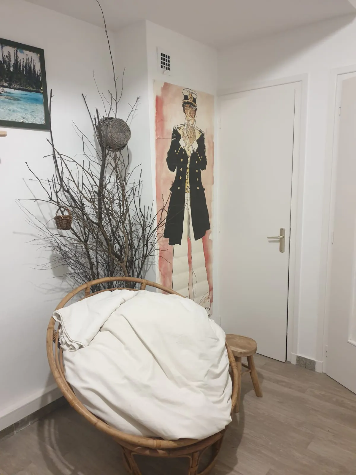Cabinet de Pierre-David MOINE, coin d'une pièce avec un fauteuil en rotin rond avec un coussin blanc, un tabouret en bois, un tableau d'une personne en uniforme militaire sur le mur et des branches décoratives avec des paniers suspendus.