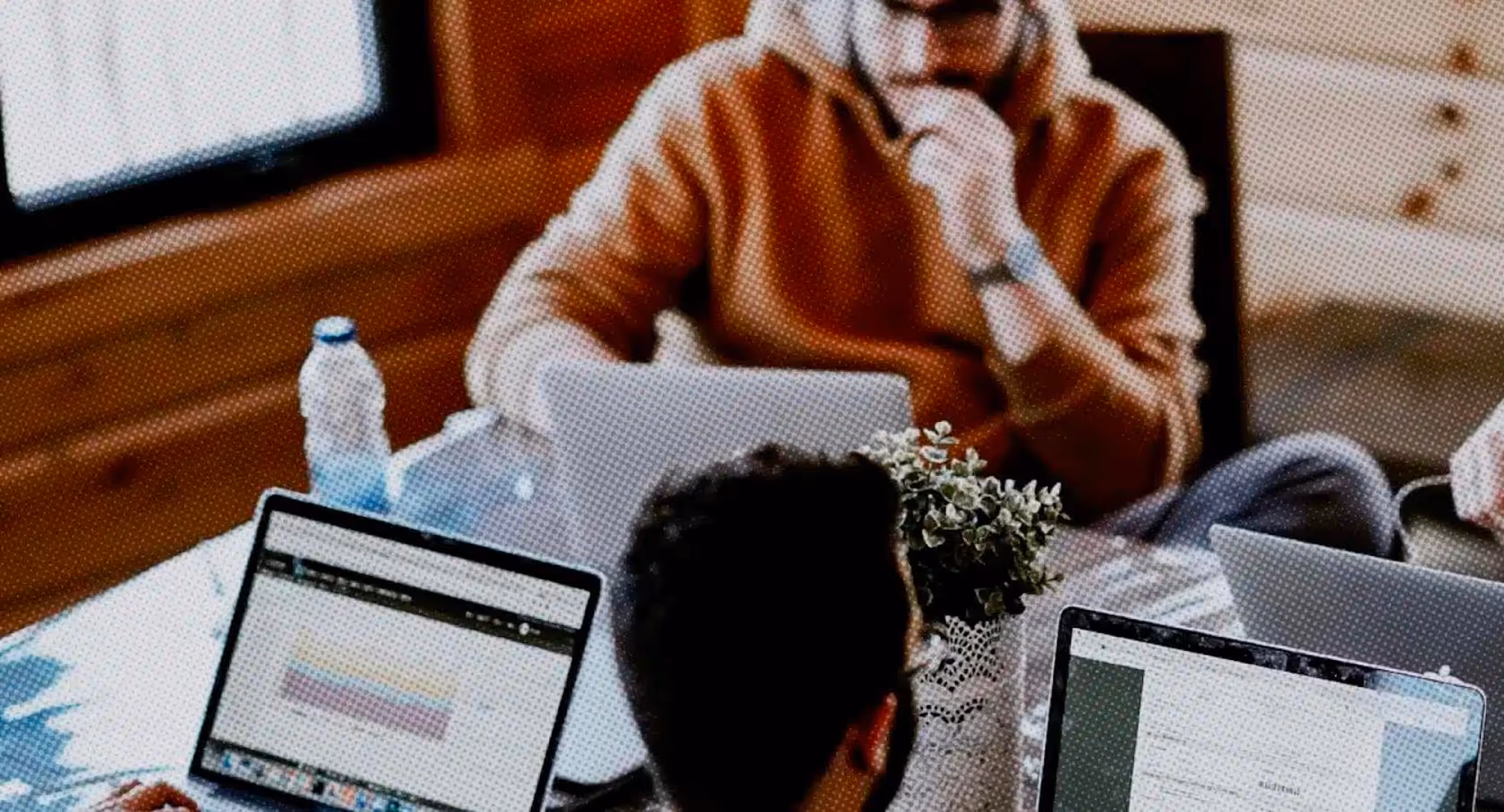 Two people working on laptops at a wooden table with a water bottle and a small potted plant.