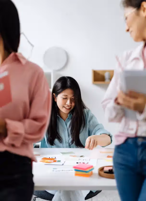 Young woman in a light blue shirt working with colorful sticky notes at a white table, while two blurred people stand on the sides.