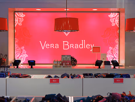 Vera Bradley store display with a bright pink background, patterned edges, hanging red lamps, and shelves filled with folded merchandise.