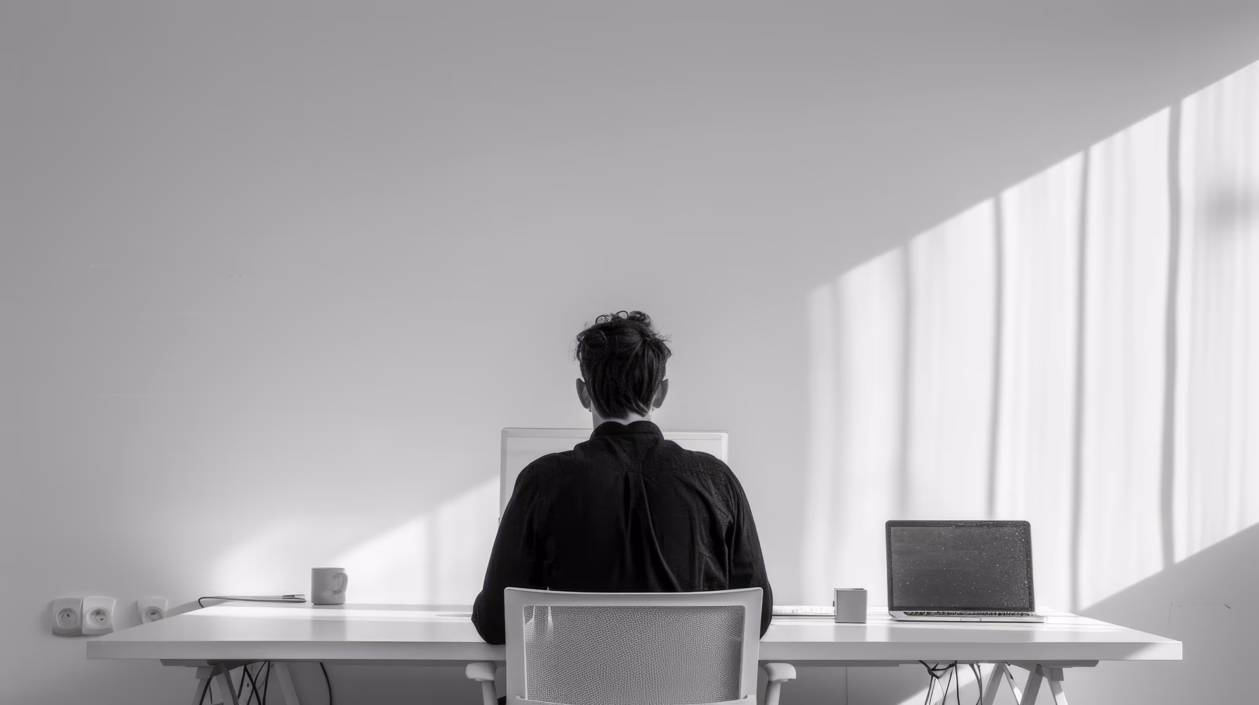 Person wearing a black shirt sitting at a white desk with two laptops in a minimalist room lit by sunlight through a window.