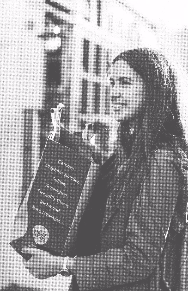 Young woman smiling and holding a Whole Foods paper shopping bag with London neighborhood names printed on it.
