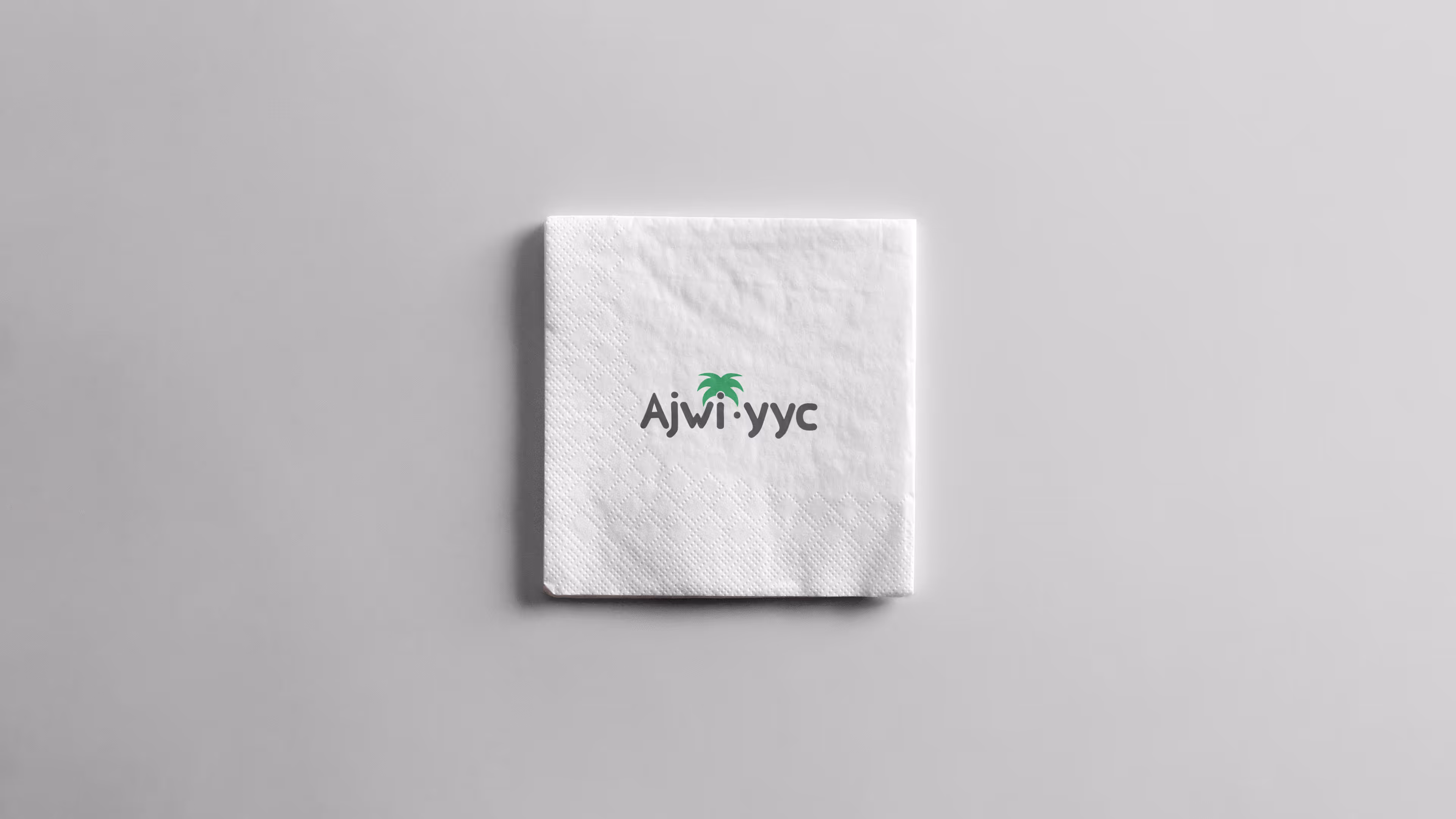 White napkin featuring the Ajwi yyc logo with a palm tree integrated into the letter 'i', placed on a light beige background. The napkin has a subtle textured pattern along one edge.