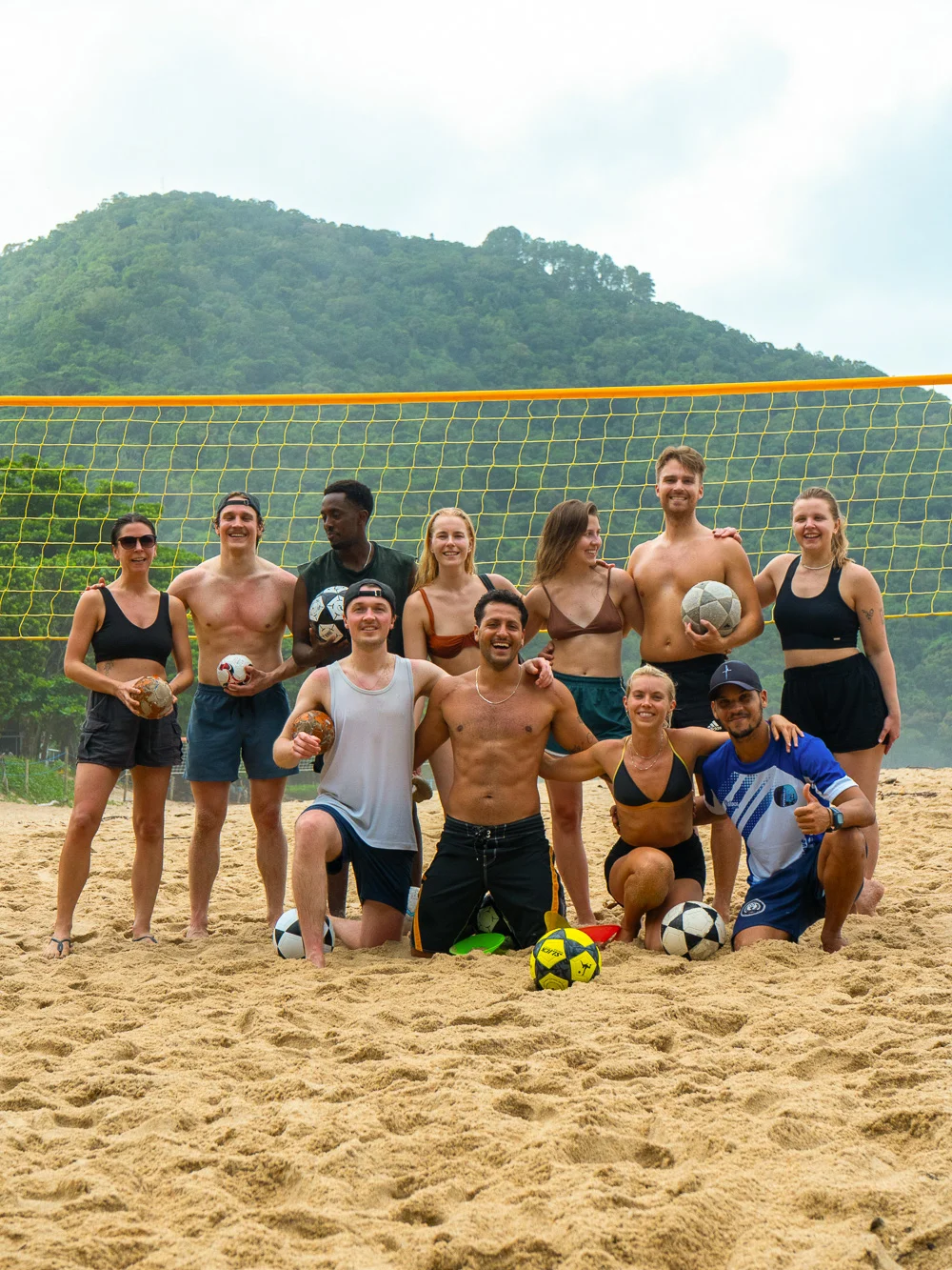 Big group photo on the beach holding footballs, green hill behind them