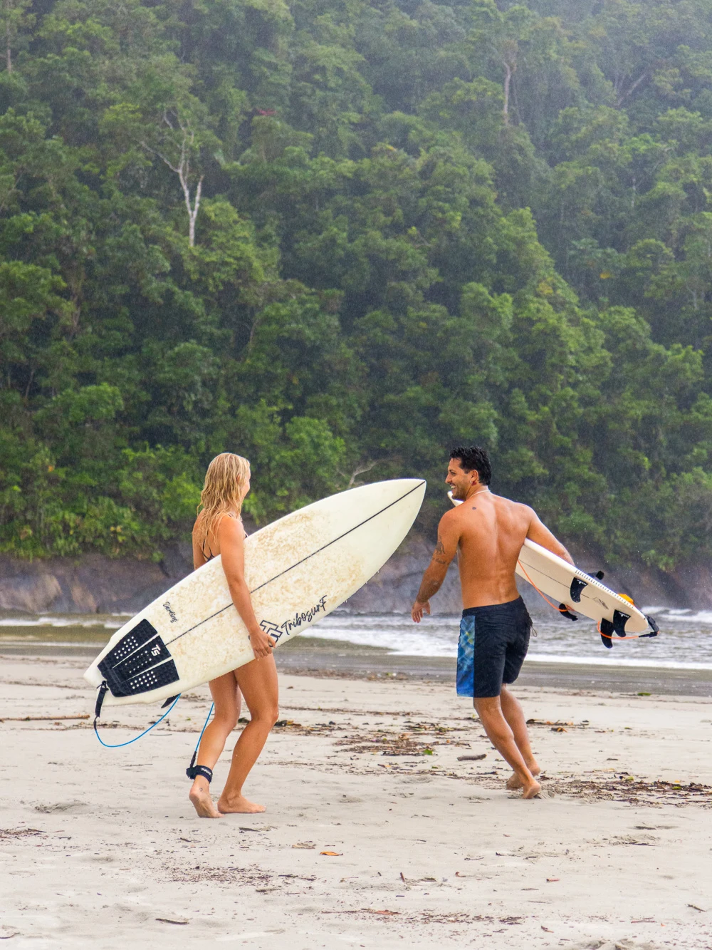 Two surfers carrying boards up the beach, laughing, jungle behind them