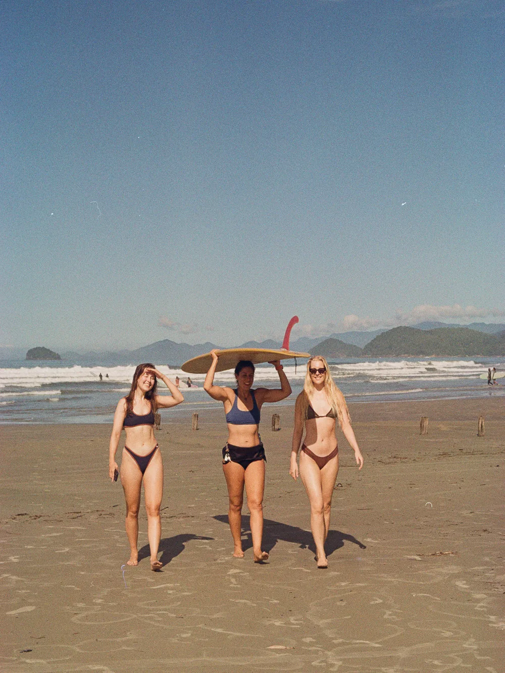 Three women walking on the beach, one carrying a board overhead, mountains behind