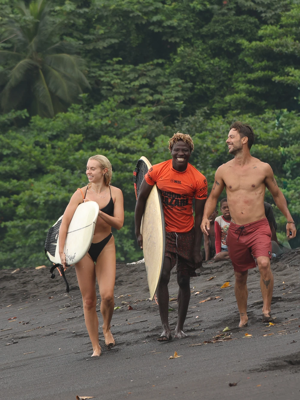 Three surfers laughing while carrying boards along a dark sand beach