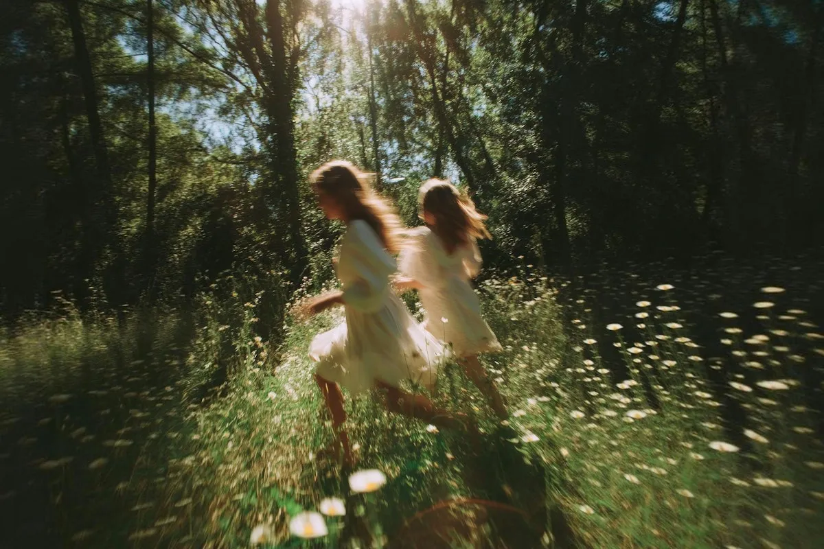 To jenter i hvite kjoler løper gjennom en solfylt, blomstereng med høye trær i bakgrunnen.
