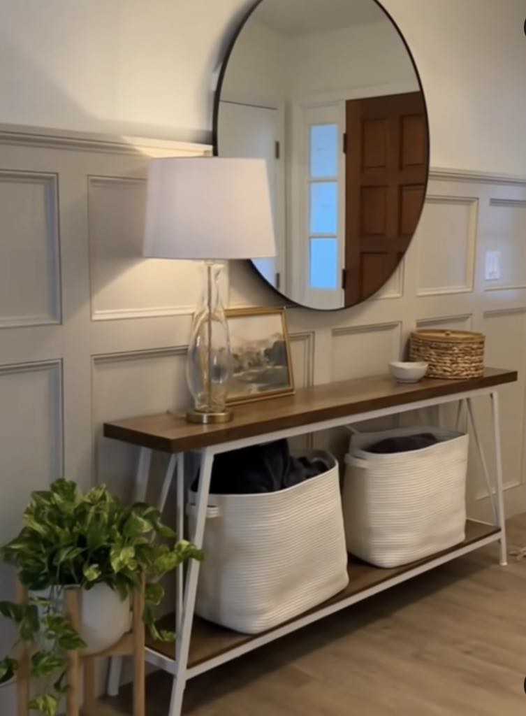 The photo shows a small hallway table with a wooden countertop. A big circle mirror is above it. The wall is a nice white with a plant in front.