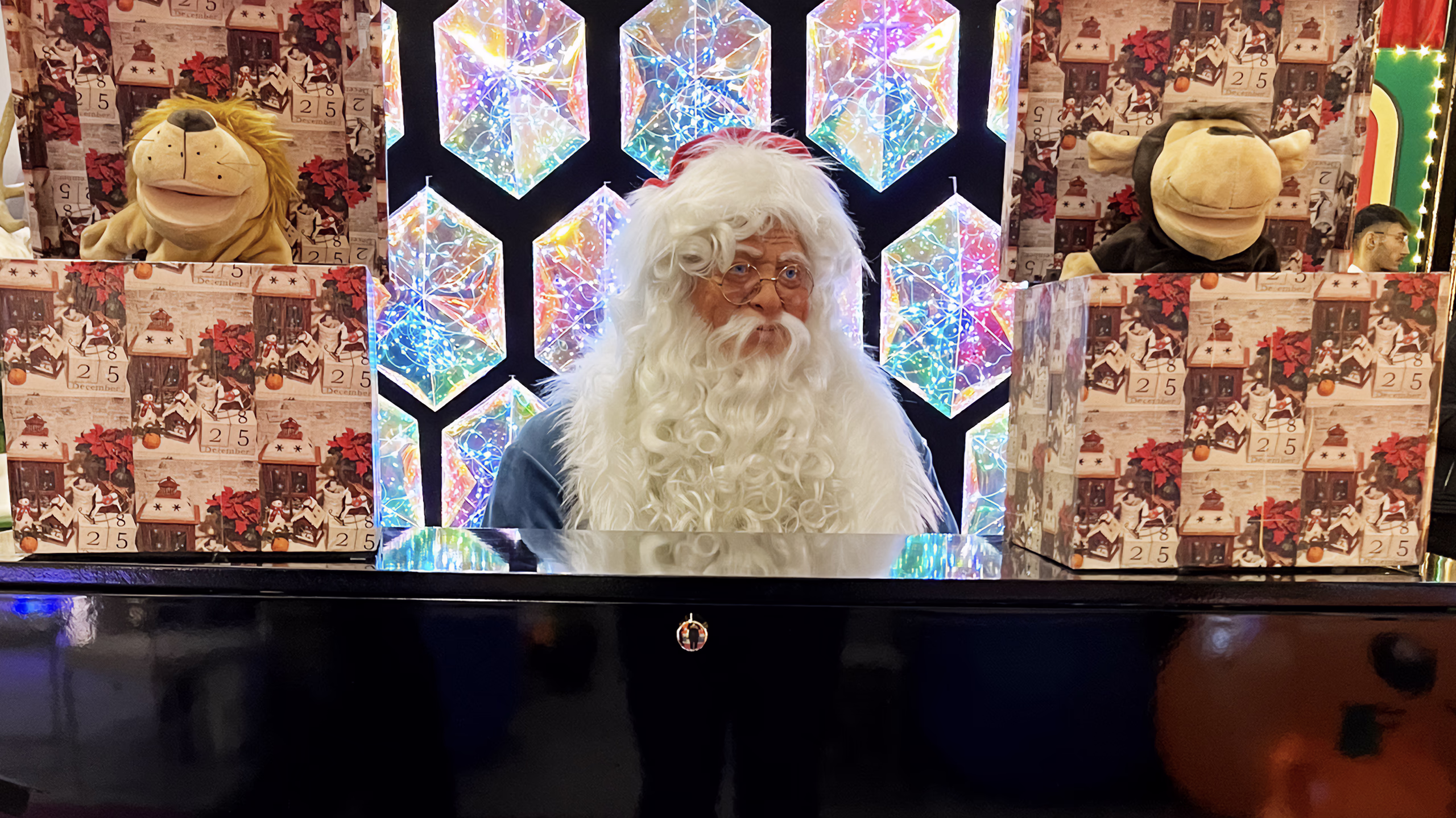 Santa Claus with white beard and glasses behind a dark counter, multicolored hexagonal luminous panels in the background, numbered Advent calendars and animated characters on the sides