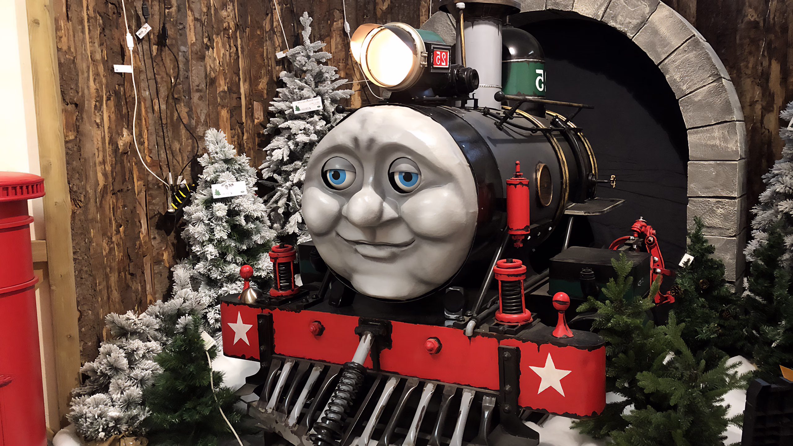 Black animatronic locomotive with smiling face and blue eyes, red details with white stars, beacon on. Arched stone tunnels, snowy Christmas trees and red lanterns as decorations