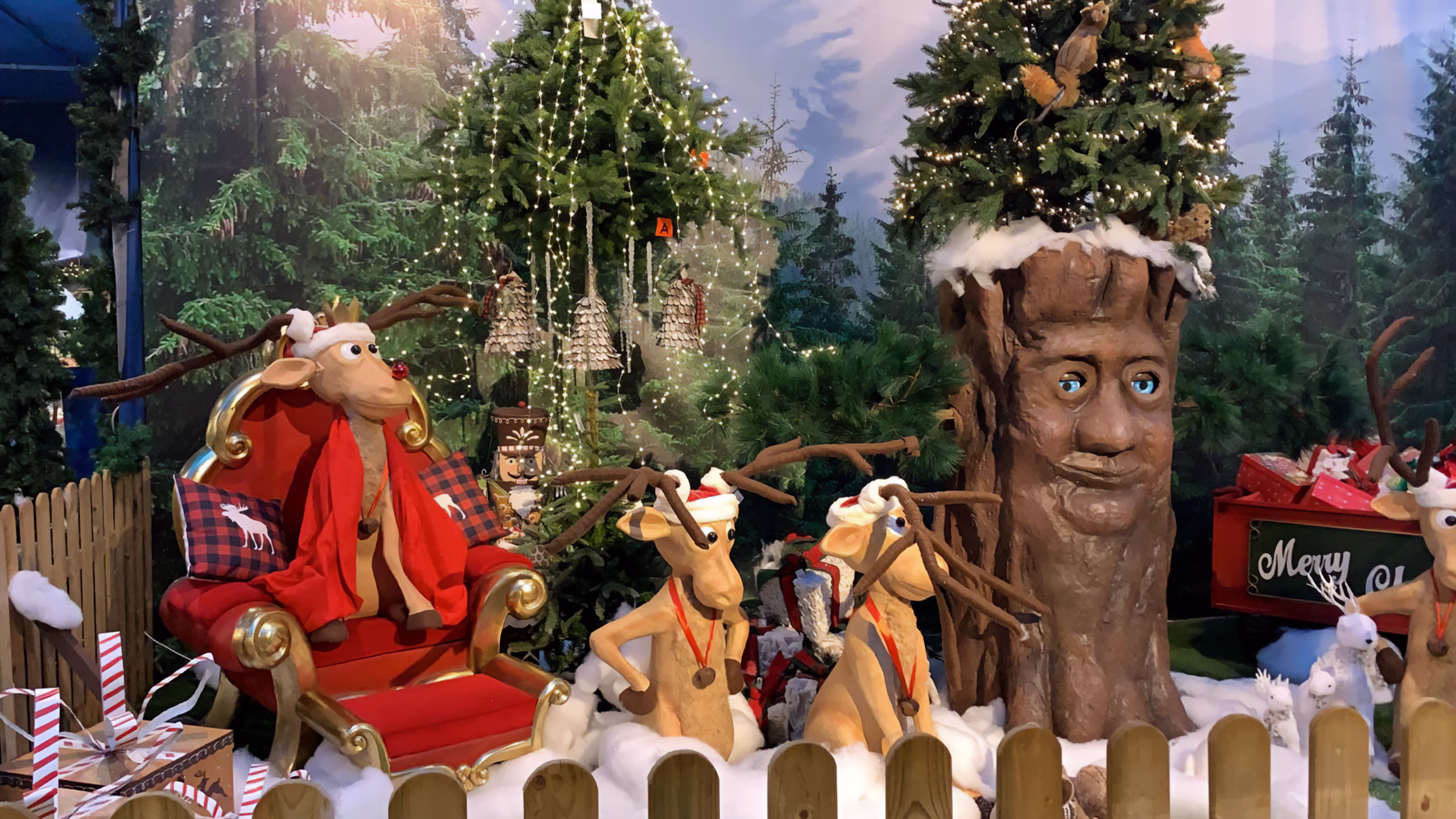Christmas scene with a reindeer in a red plaid jacket sitting on a golden sleigh, two reindeer with horns in front of the sleigh, animatronic tree with a human face and a green crown. Background with illuminated fir trees, artificial snow and wooden fence